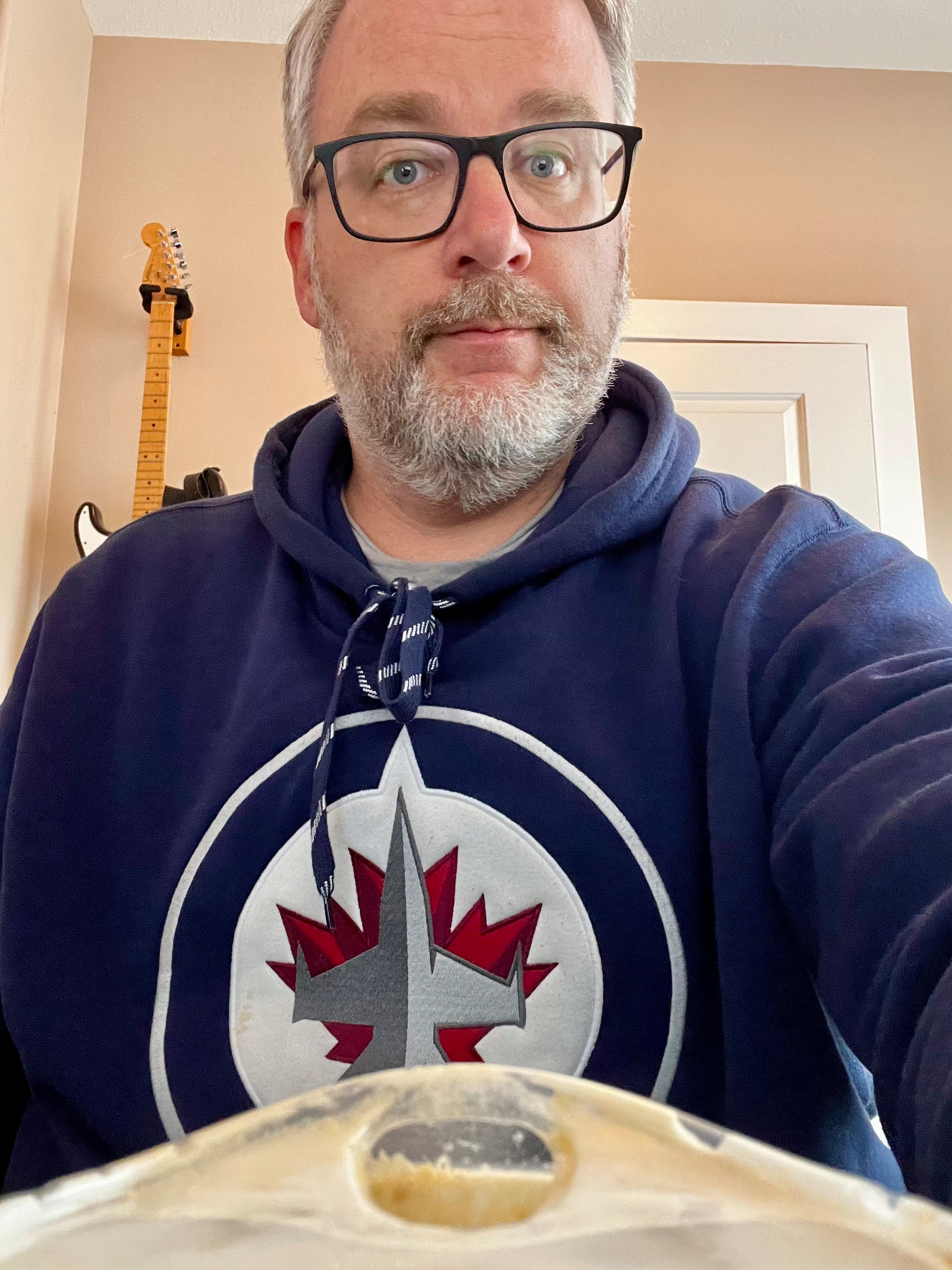A person (me!) wearing glasses and a navy hoodie with a winnipg jets emblem is sitting indoors with a iced coffee lid at the bottom 