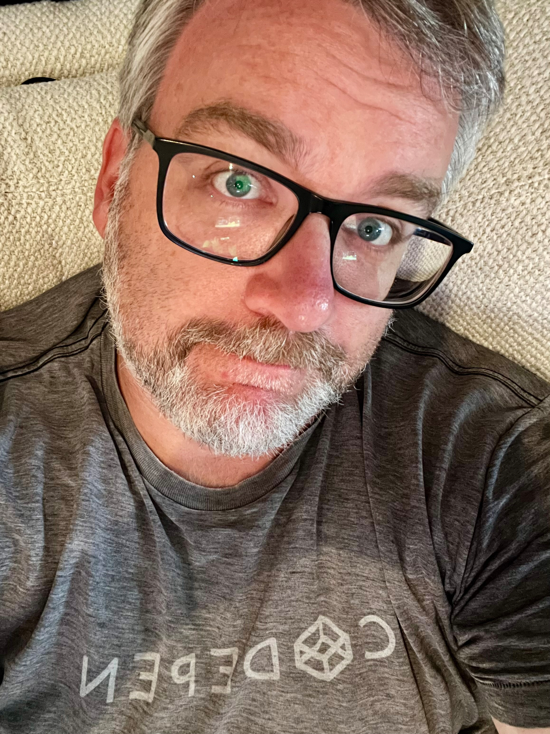 A man with gray hair, a beard, and black-framed glasses is looking up at the camera while lying on a light-colored couch and wearing a gray CodePen t-shirt.