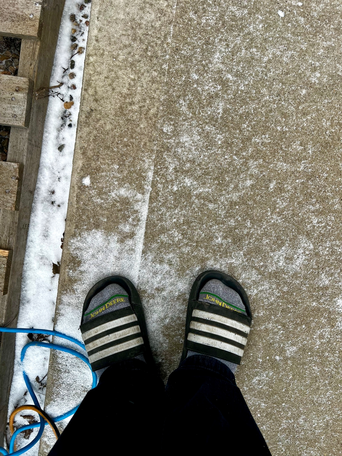 A person wearing black slides with white stripes stands on a concrete surface partially covered with snow near a wooden structure and a blue cable.