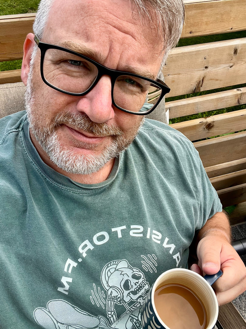 A man with glasses and a beard is sitting outdoors, holding a cup of coffee and wearing a green T-shirt with a graphic design.