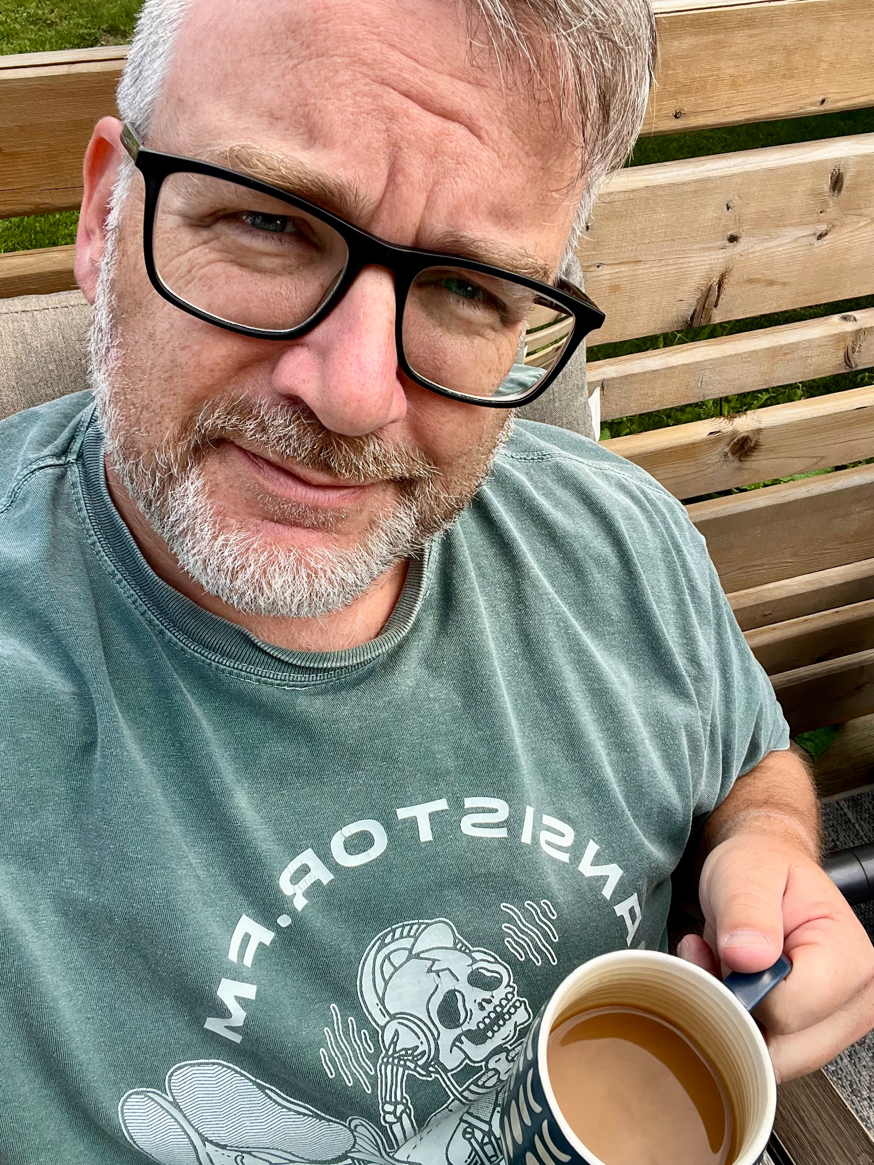 A man with glasses and a beard is sitting outdoors, holding a cup of coffee and wearing a green T-shirt with a graphic design.
