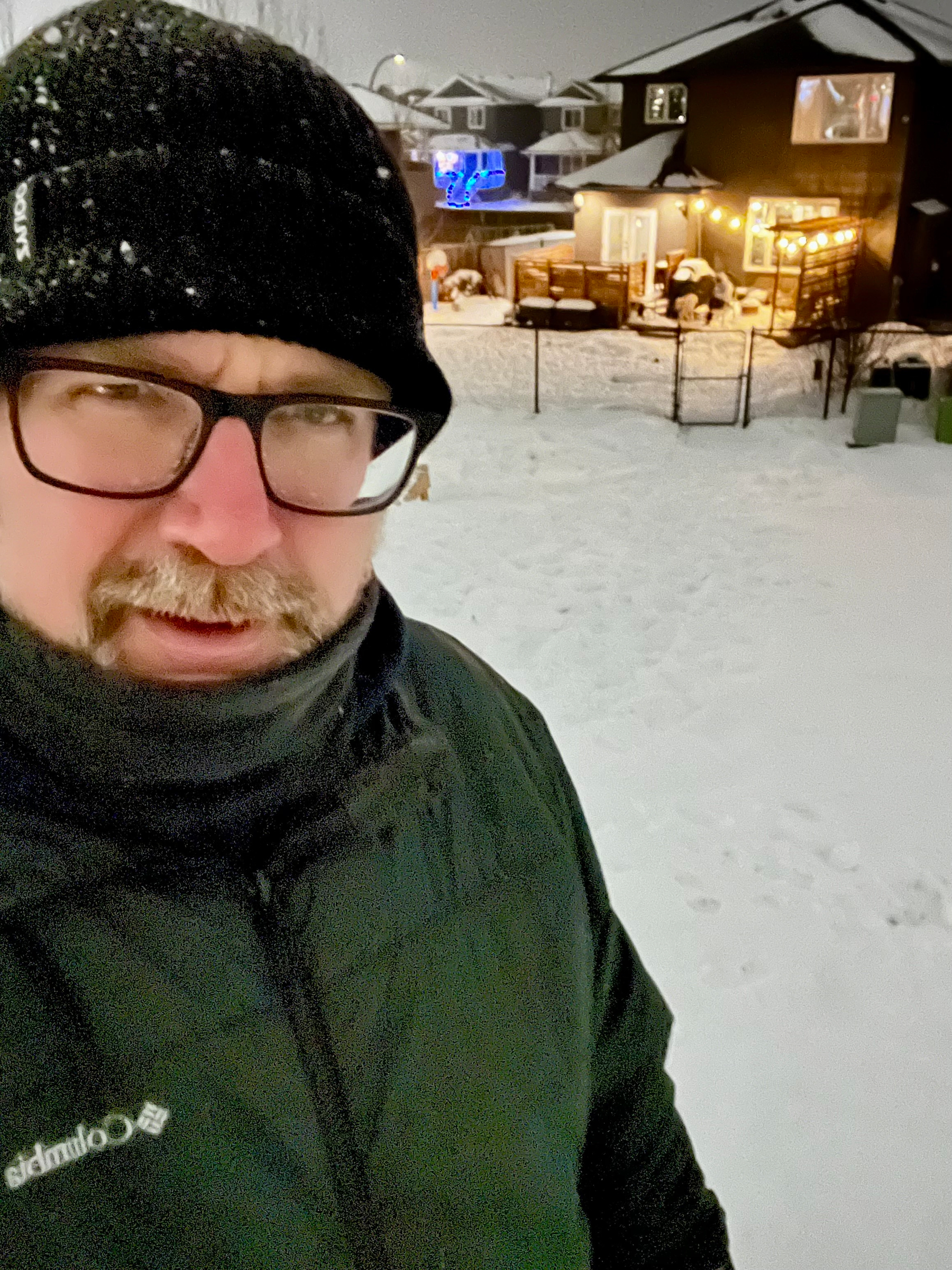 A person wearing winter clothing stands in a snowy backyard with a house and festive lights in the background.