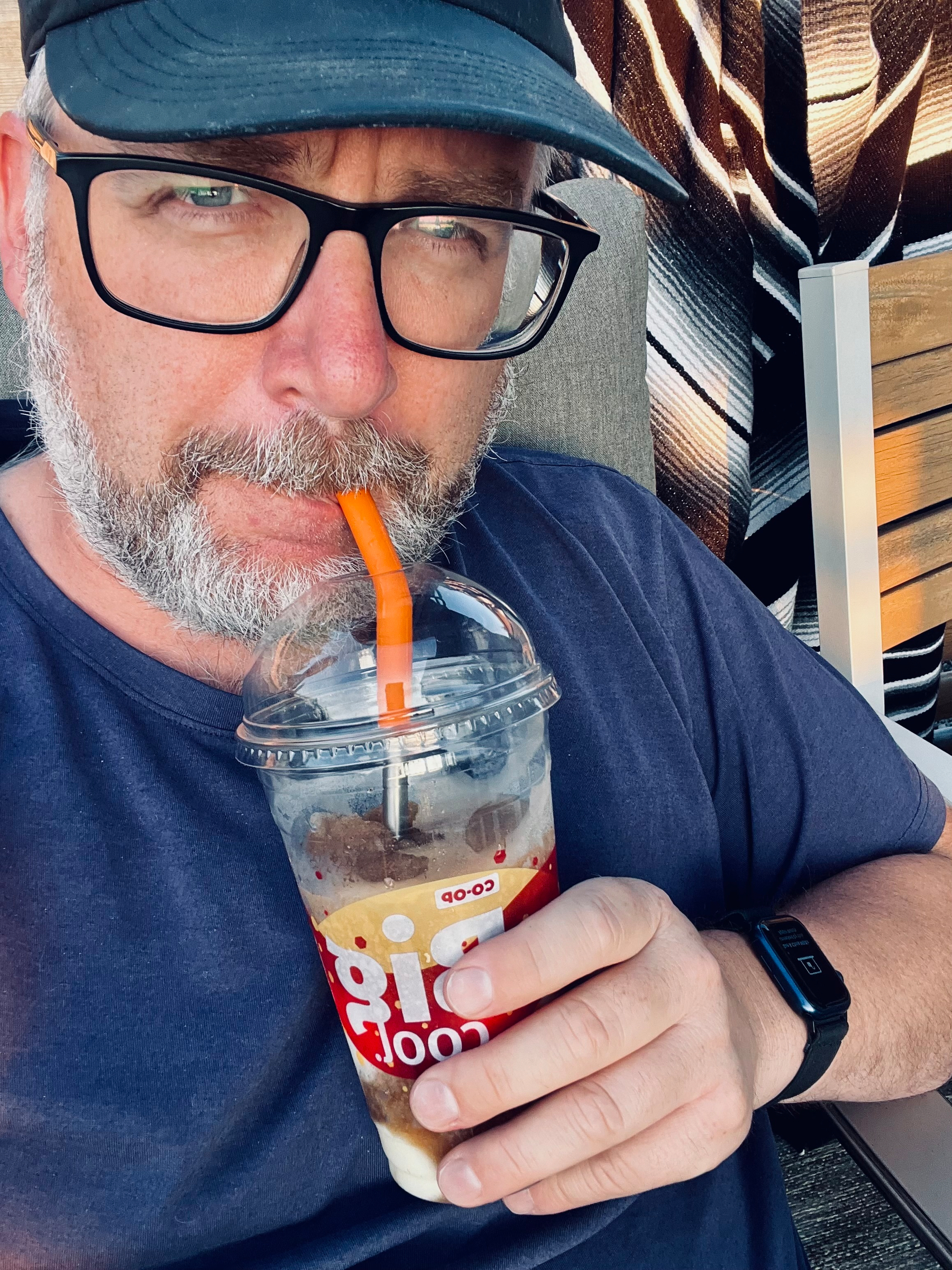 A man wearing glasses and a cap is sipping a slurpee through an orange straw from a plastic cup.