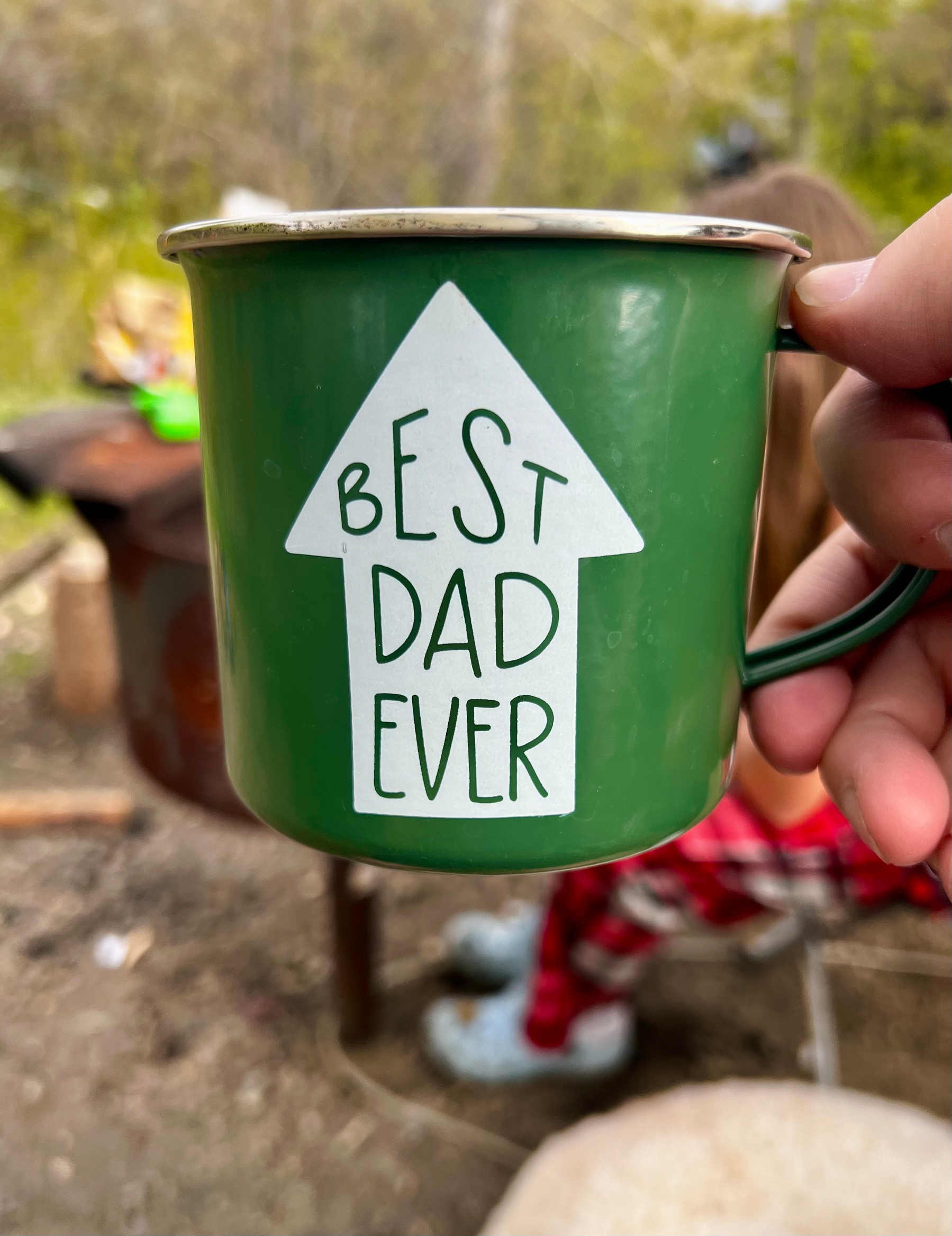 A green mug with Best Dad Ever written on it is held up outdoors, with blurred background details.