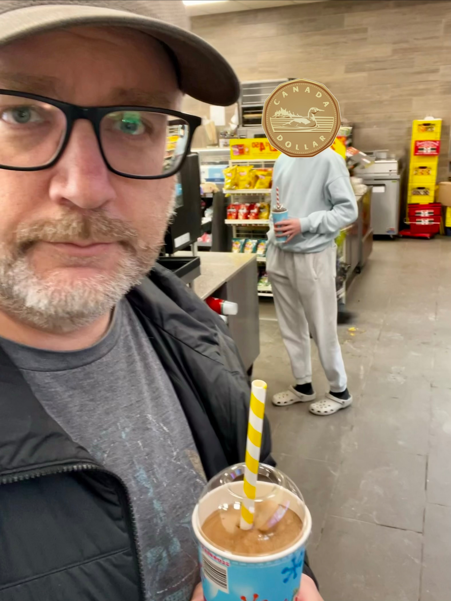 A person holding a drink with a striped straw stands in a store, while another person in the background appears to be checking their phone near a display of yellow snacks.