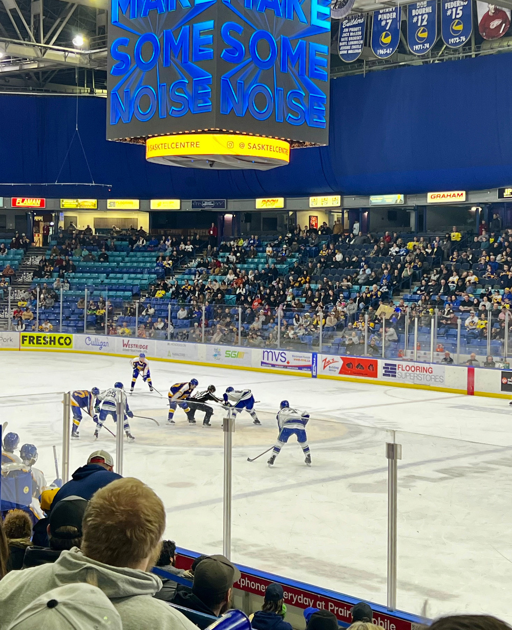 A hockey game is taking place in a crowded arena with players ready for a face-off and a scoreboard encouraging fans to make noise.