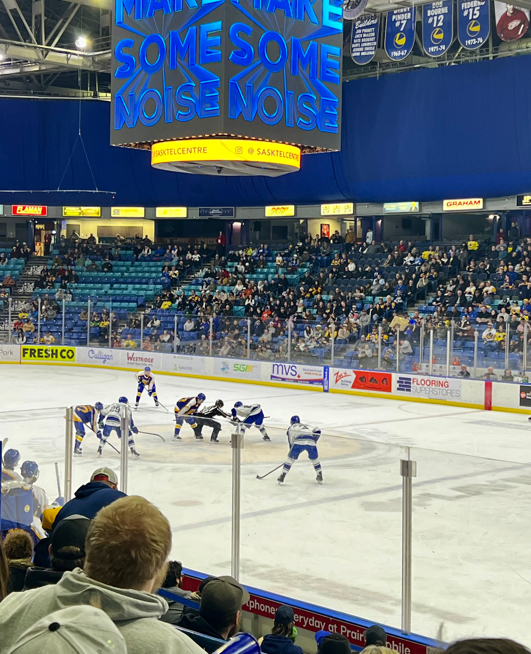 A hockey game is taking place in a crowded arena with players ready for a face-off and a scoreboard encouraging fans to make noise.