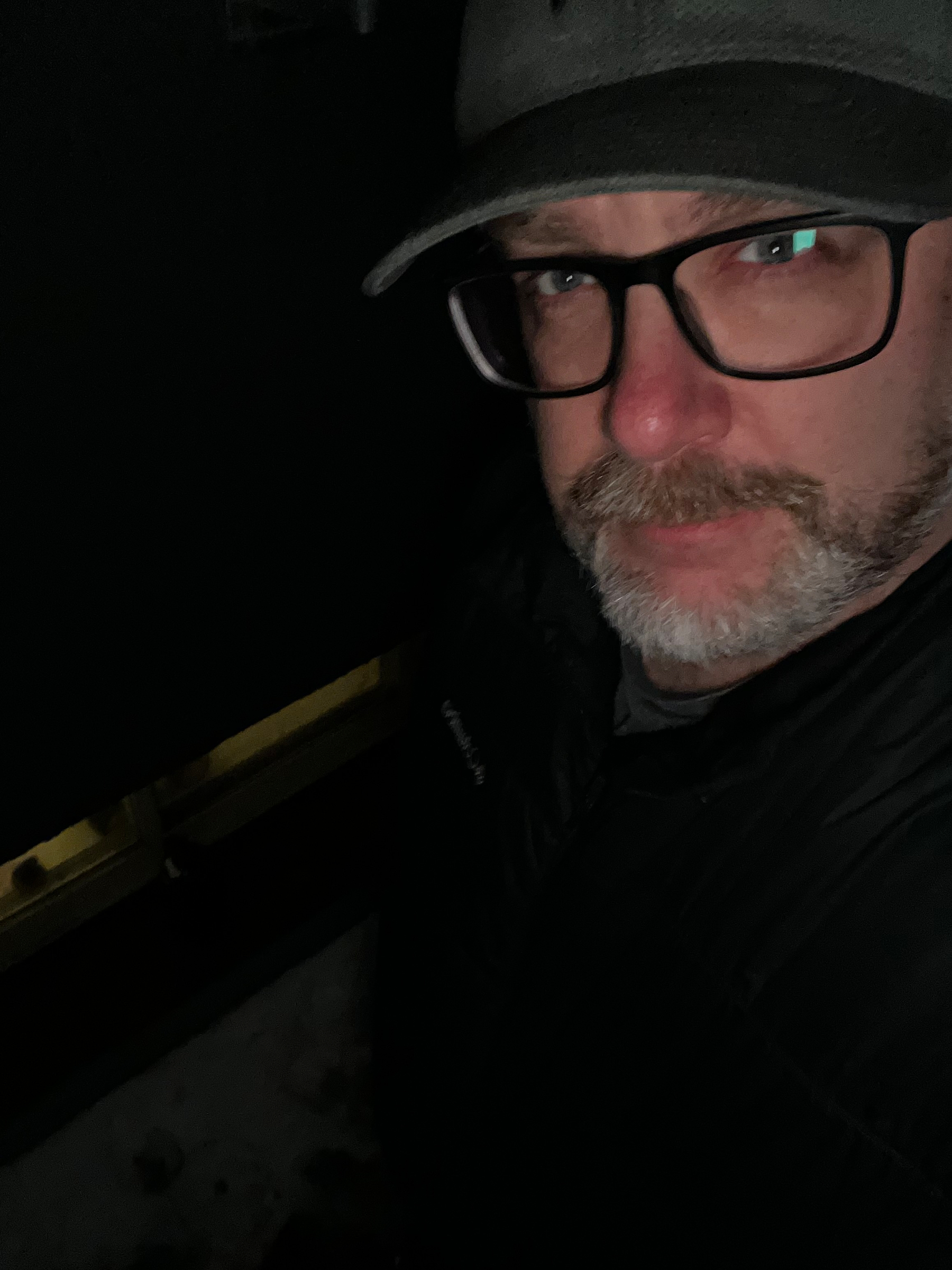 A person wearing glasses and a cap is looking at the camera in a dimly lit setting.