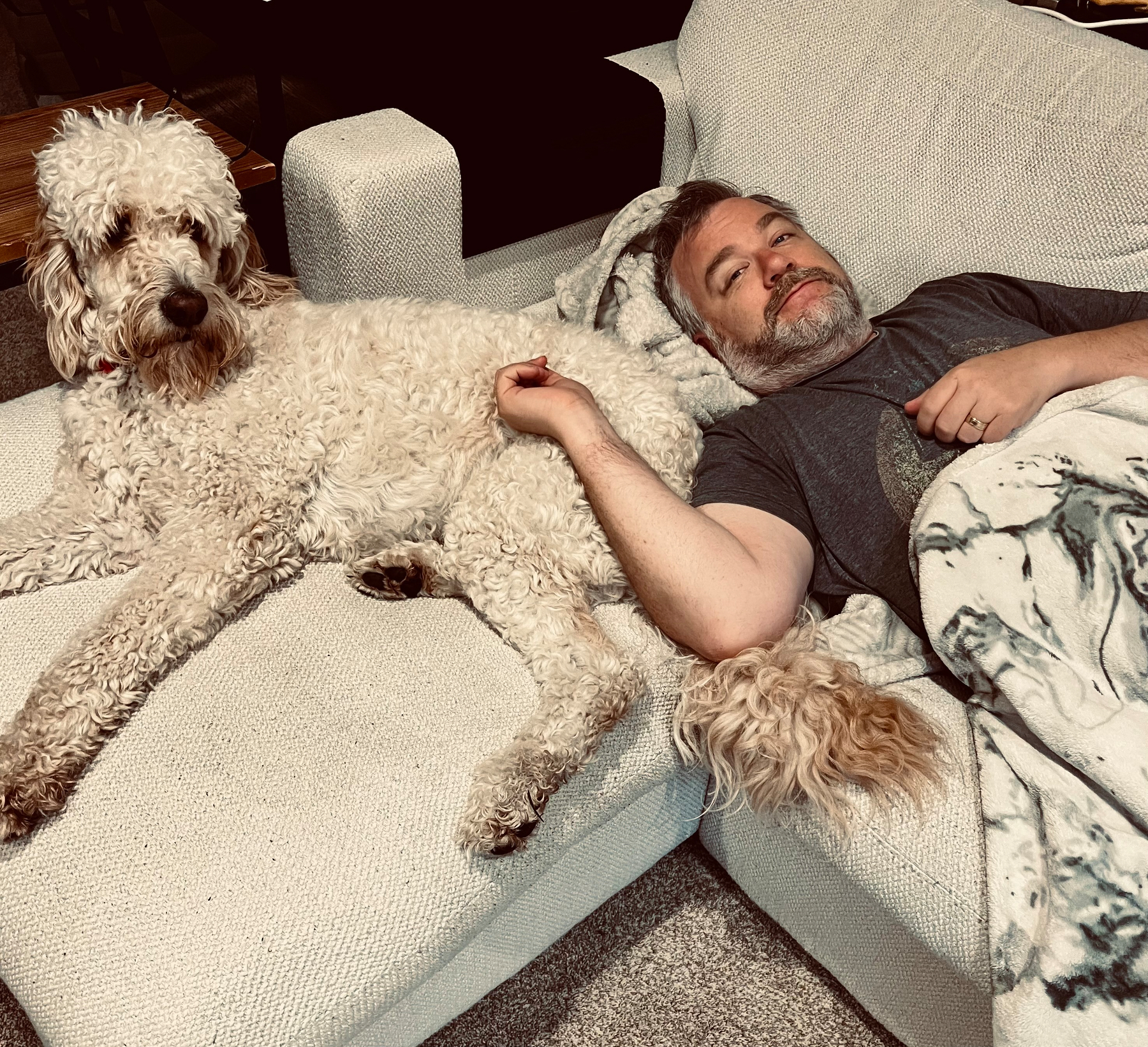 A man is lying on a light-colored sofa covered with a blanket, holding the paw of a large, curly-haired dog who is also resting on the sofa.