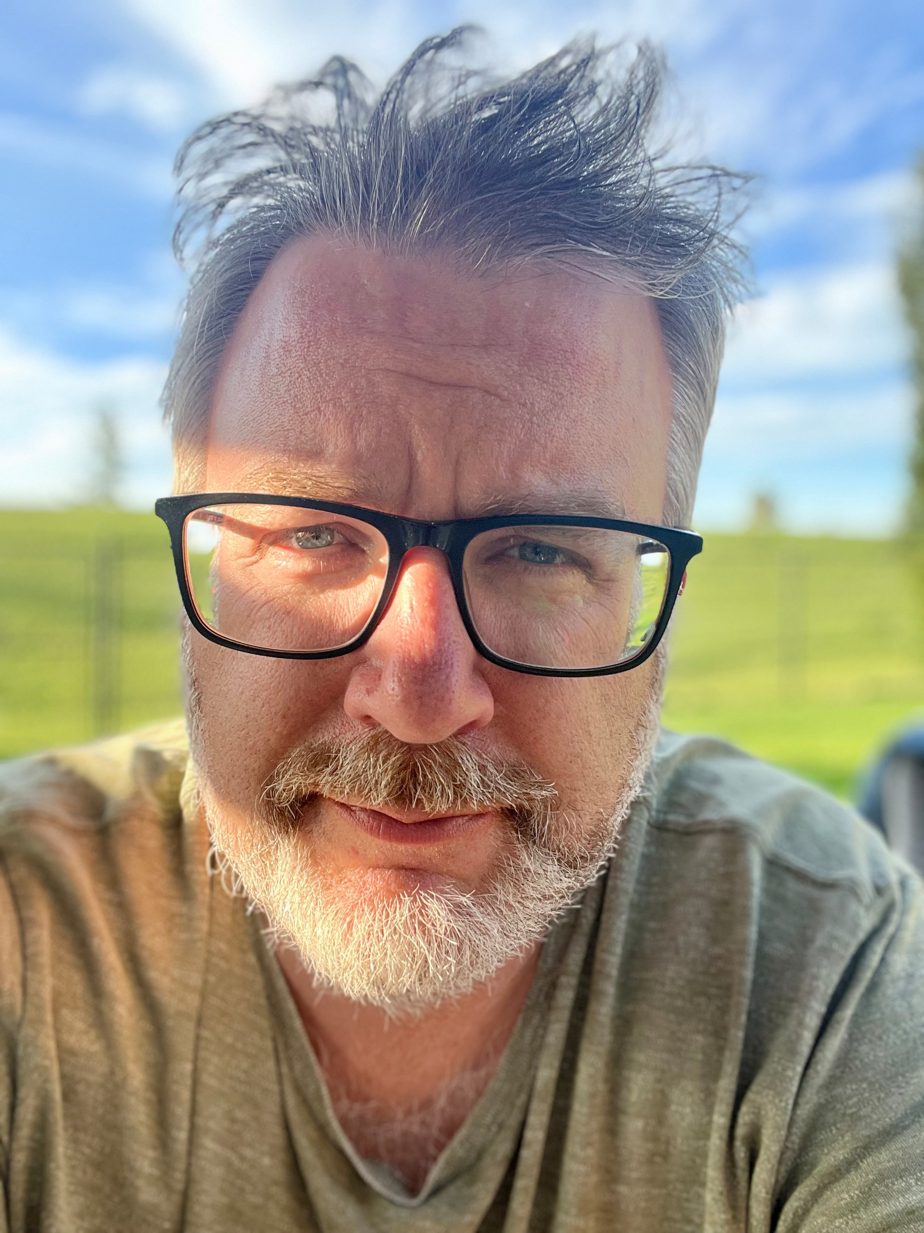 A man with glasses, a beard, and slightly tousled hair is looking directly at the camera with a green outdoor background behind him.