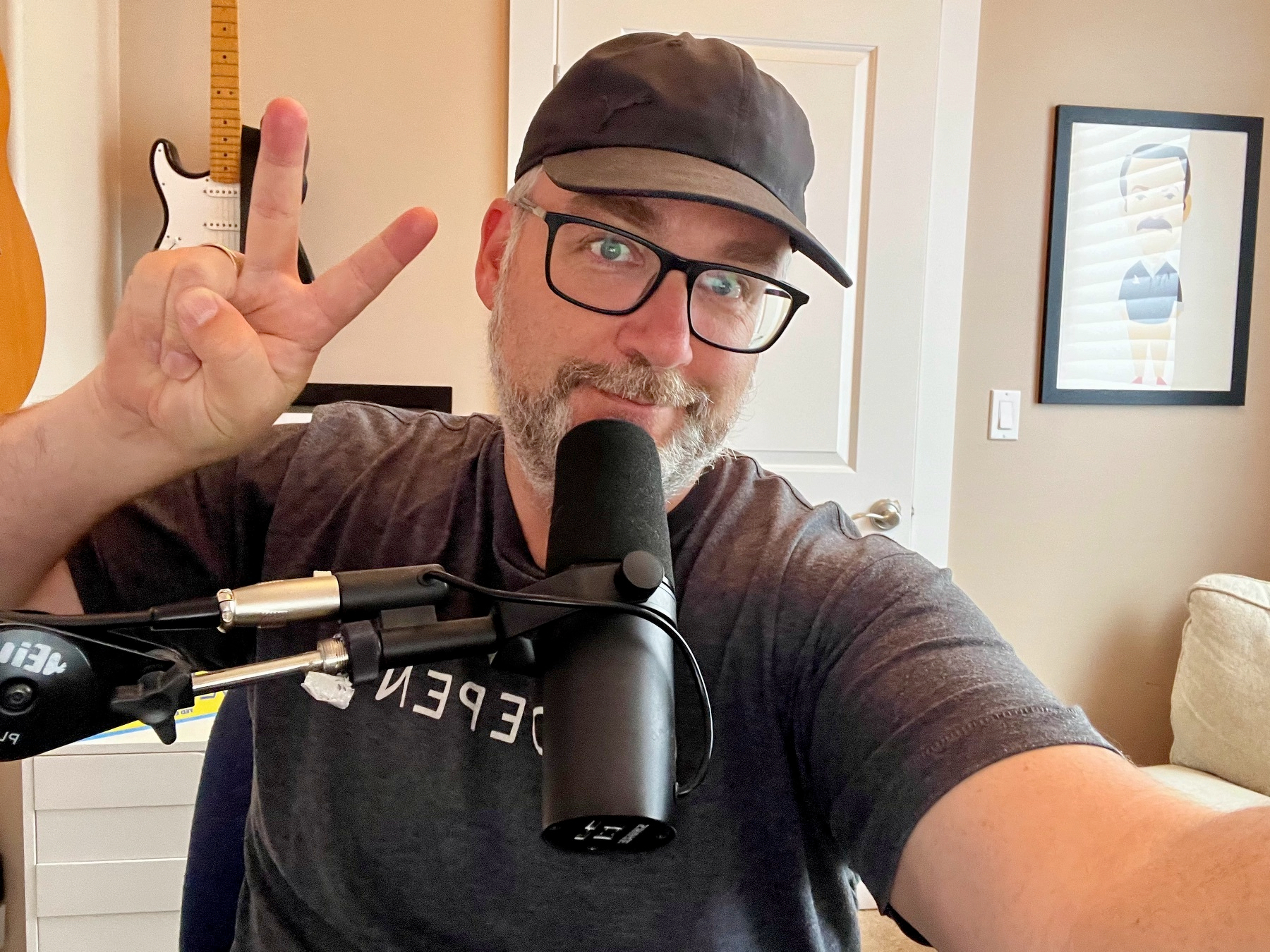 A man wearing glasses and a black cap is smiling, making a peace sign with one hand, and speaking into a microphone in a cozy room with a guitar and framed artwork in the background.