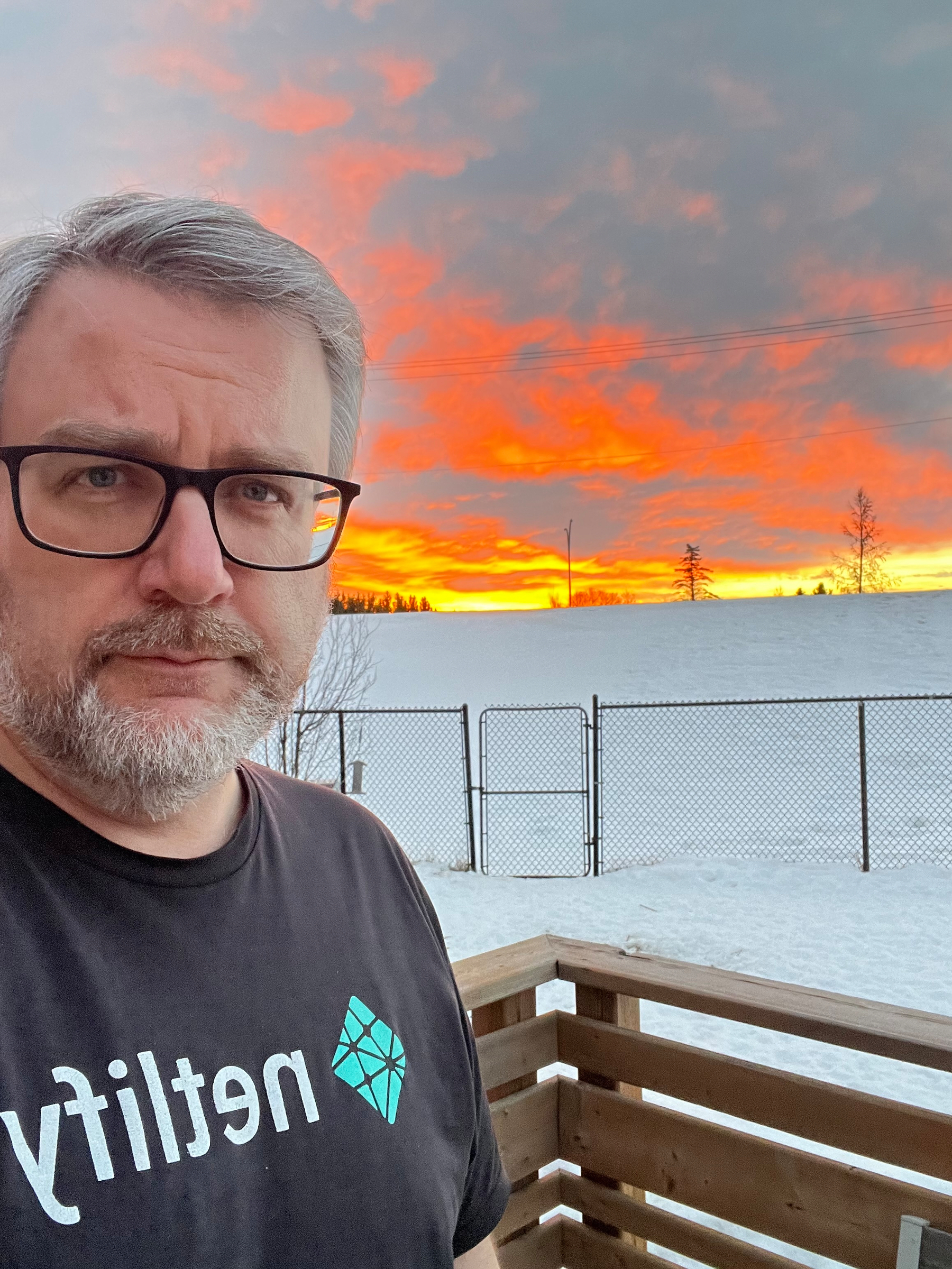 A person with glasses and a beard is standing on a snowy balcony at sunset, wearing a shirt with netlify on it.