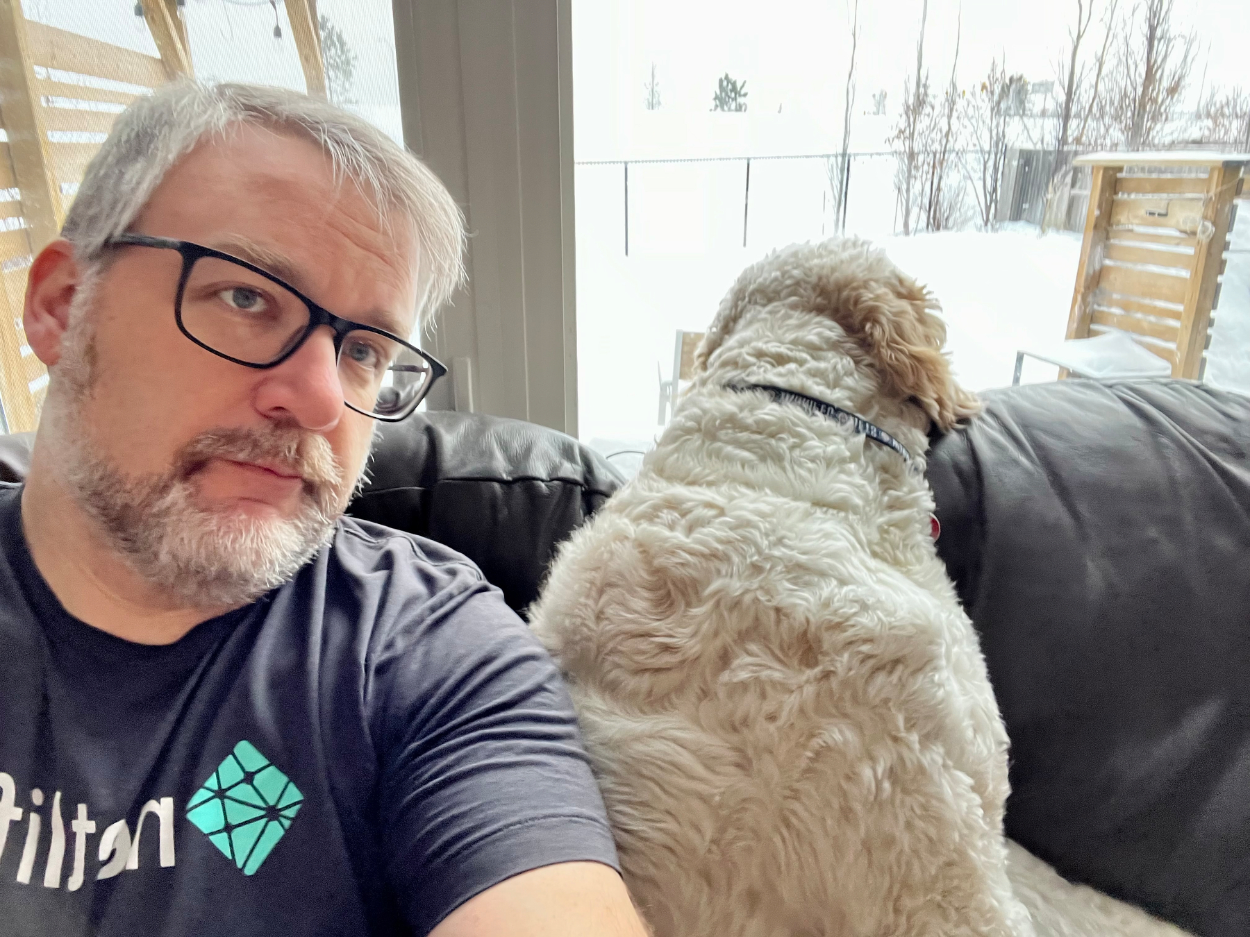 A man with glasses and a beard is sitting on a couch next to a fluffy dog looking out the window.