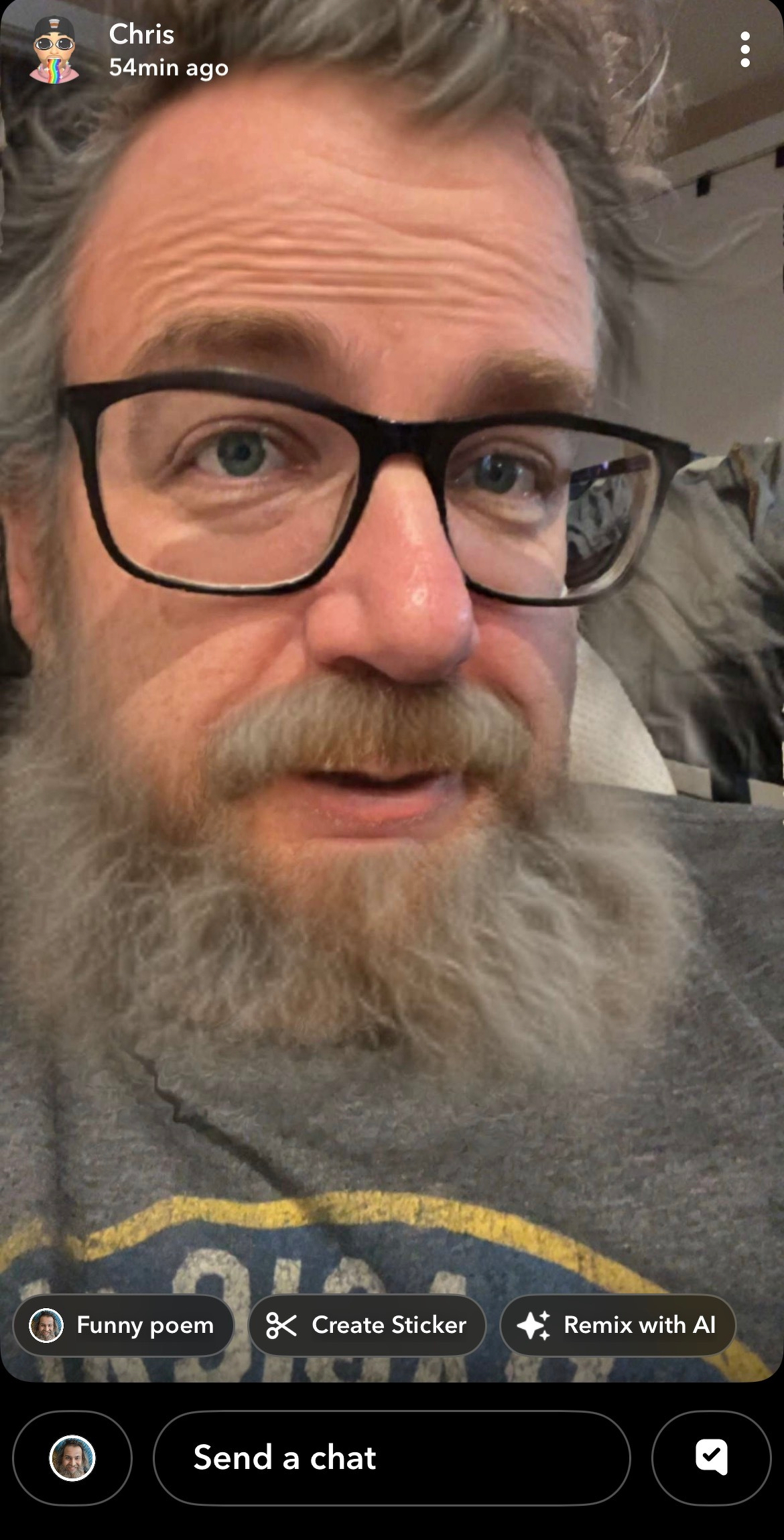 A close-up of a man with glasses, a thick gray beard, and curly hair is shown indoors.