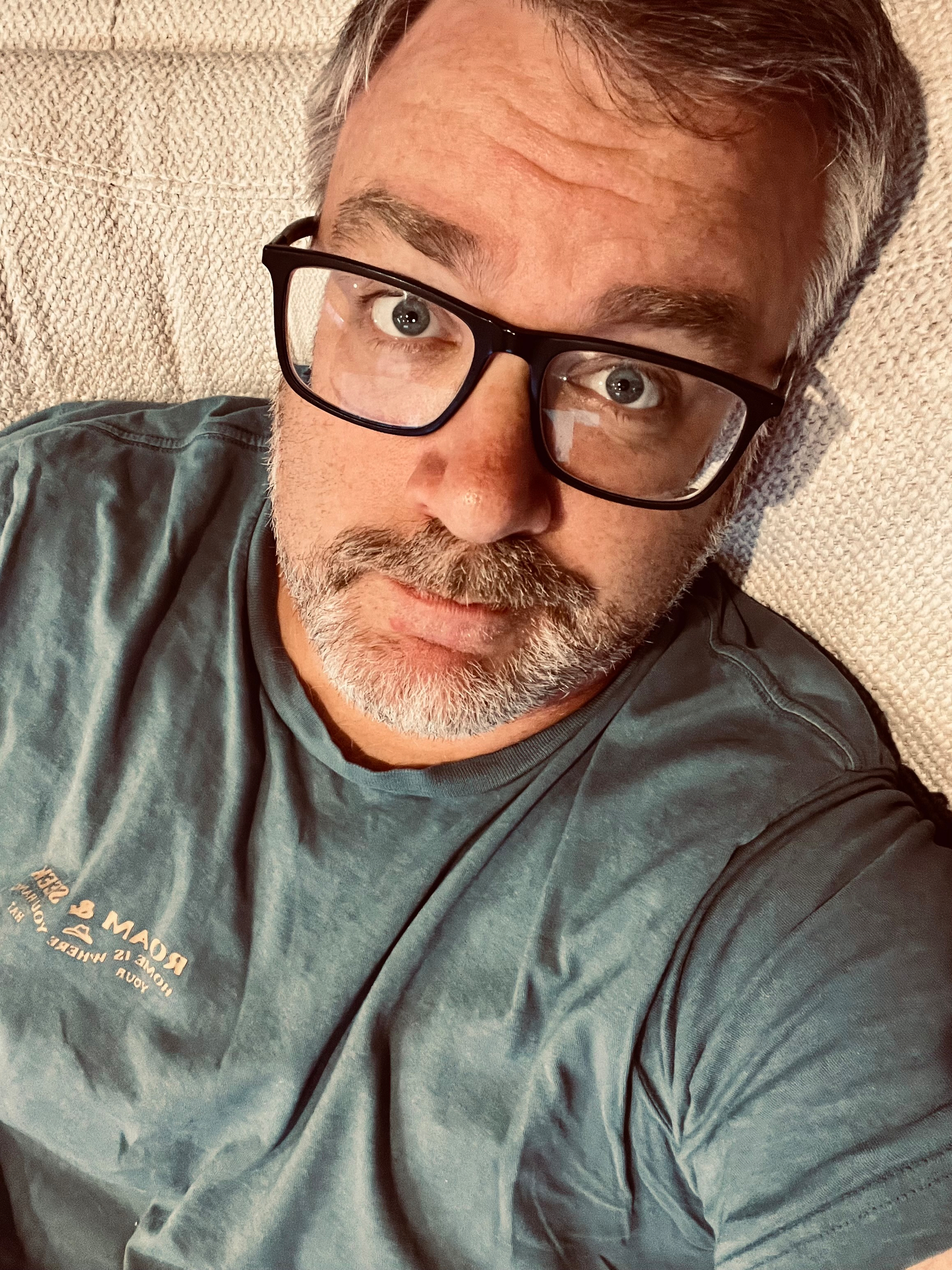 A man with glasses and a beard is lying on a textured surface, wearing a dark blue shirt.