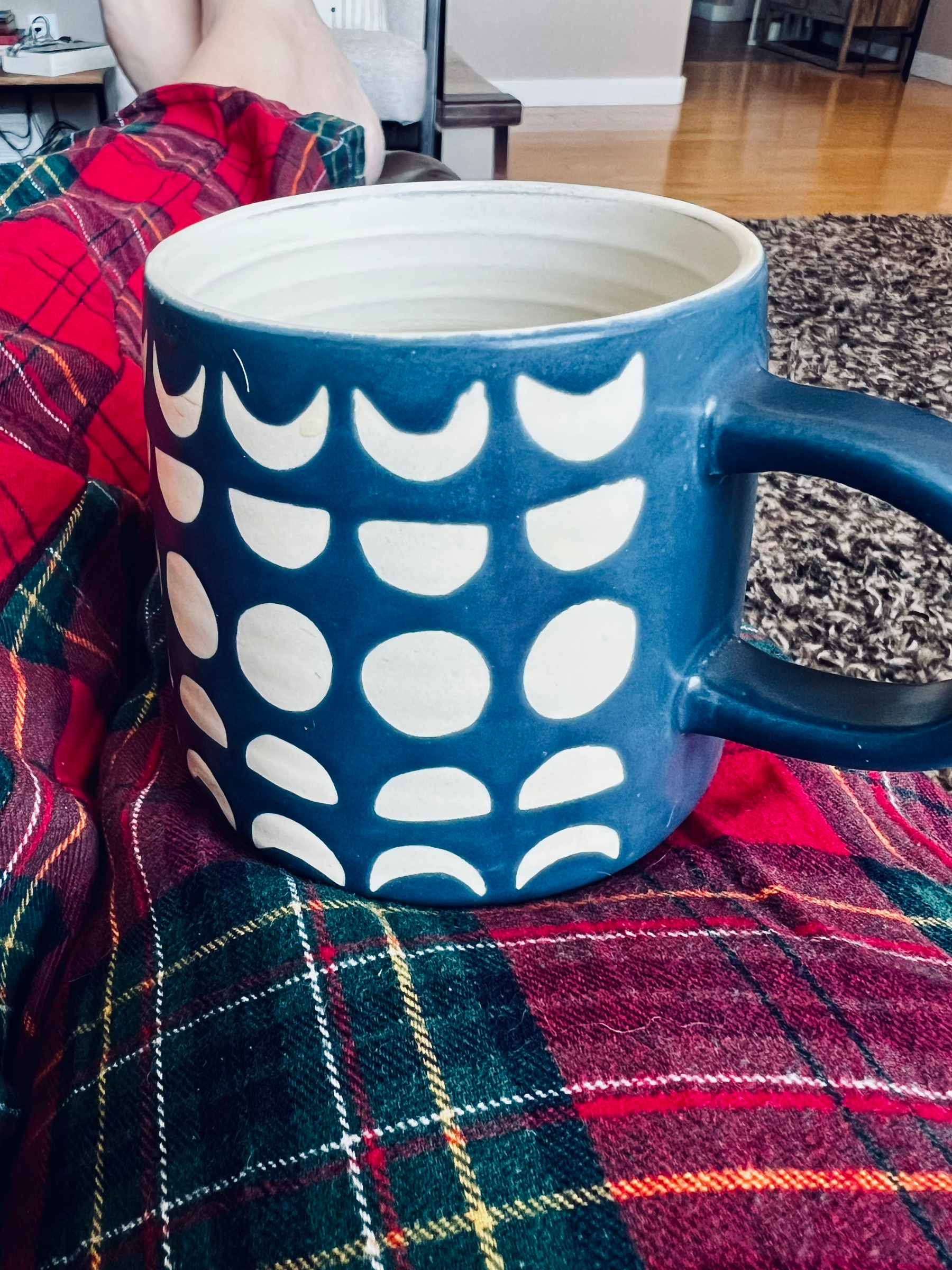 A large blue mug with a white crescent pattern is resting on someone's lap, who is wearing red plaid clothing.