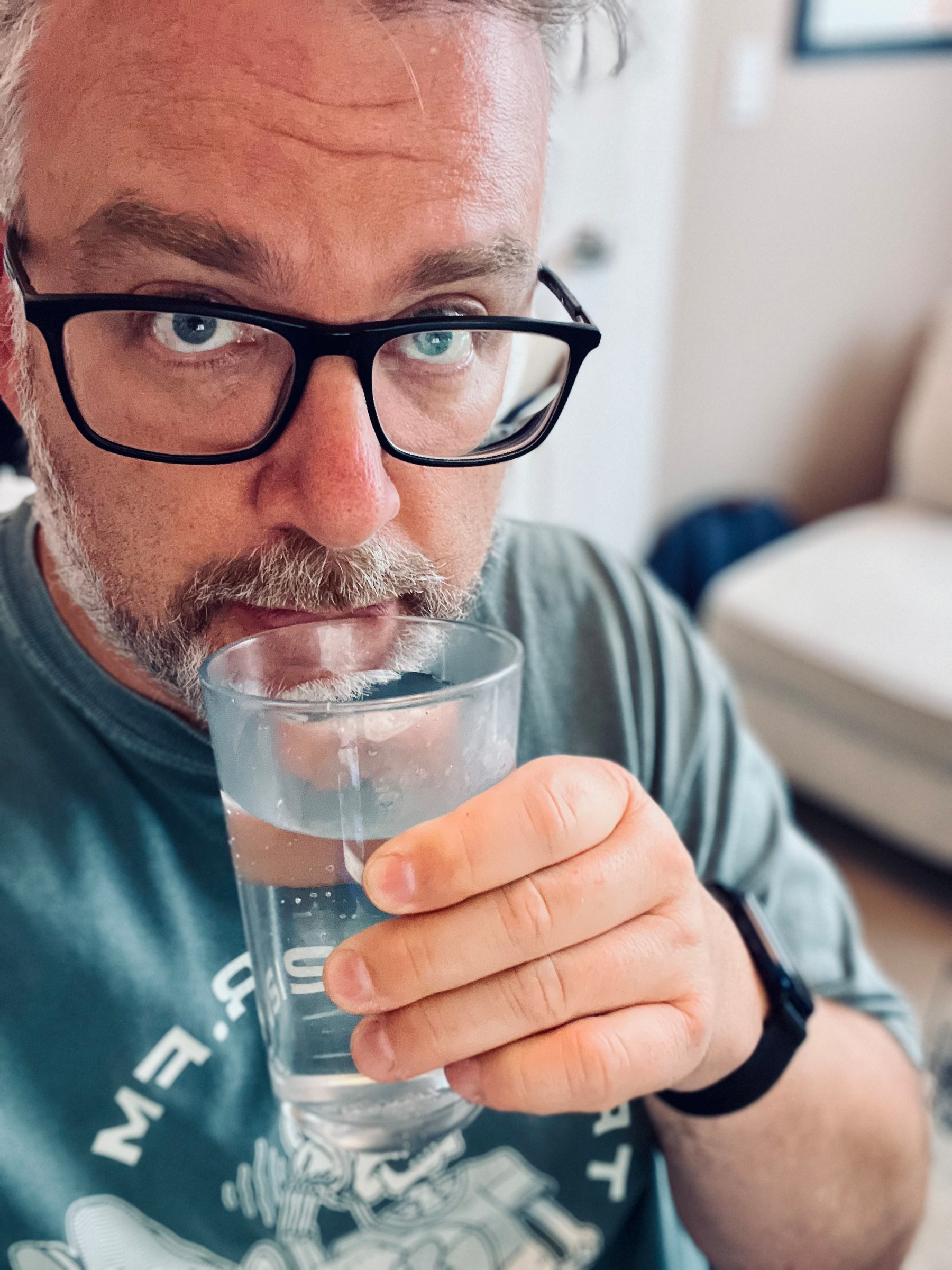 A man wearing glasses is holding and drinking from a glass of water.