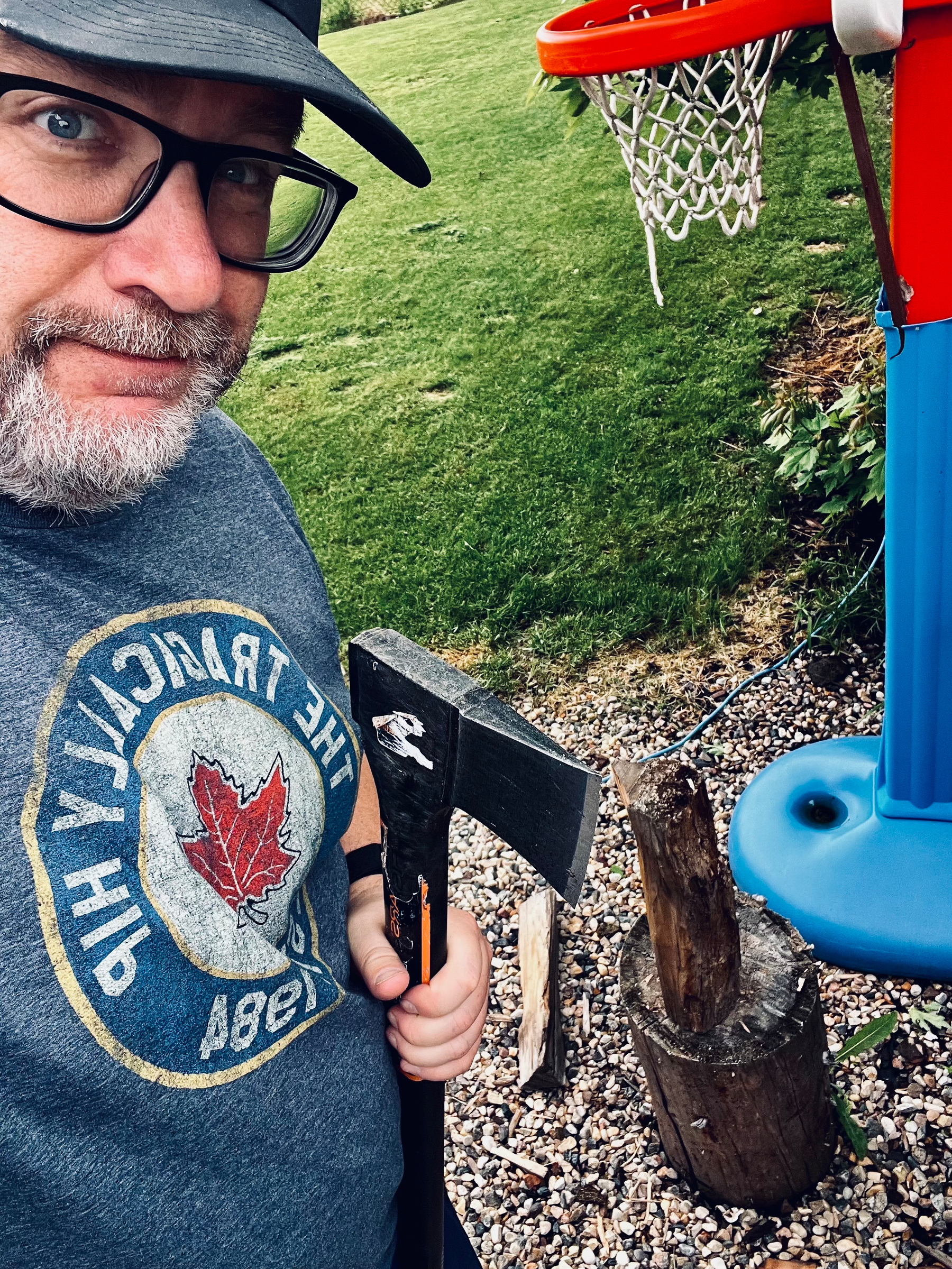 A person is holding an axe beside a tree stump near a basketball hoop in a backyard.