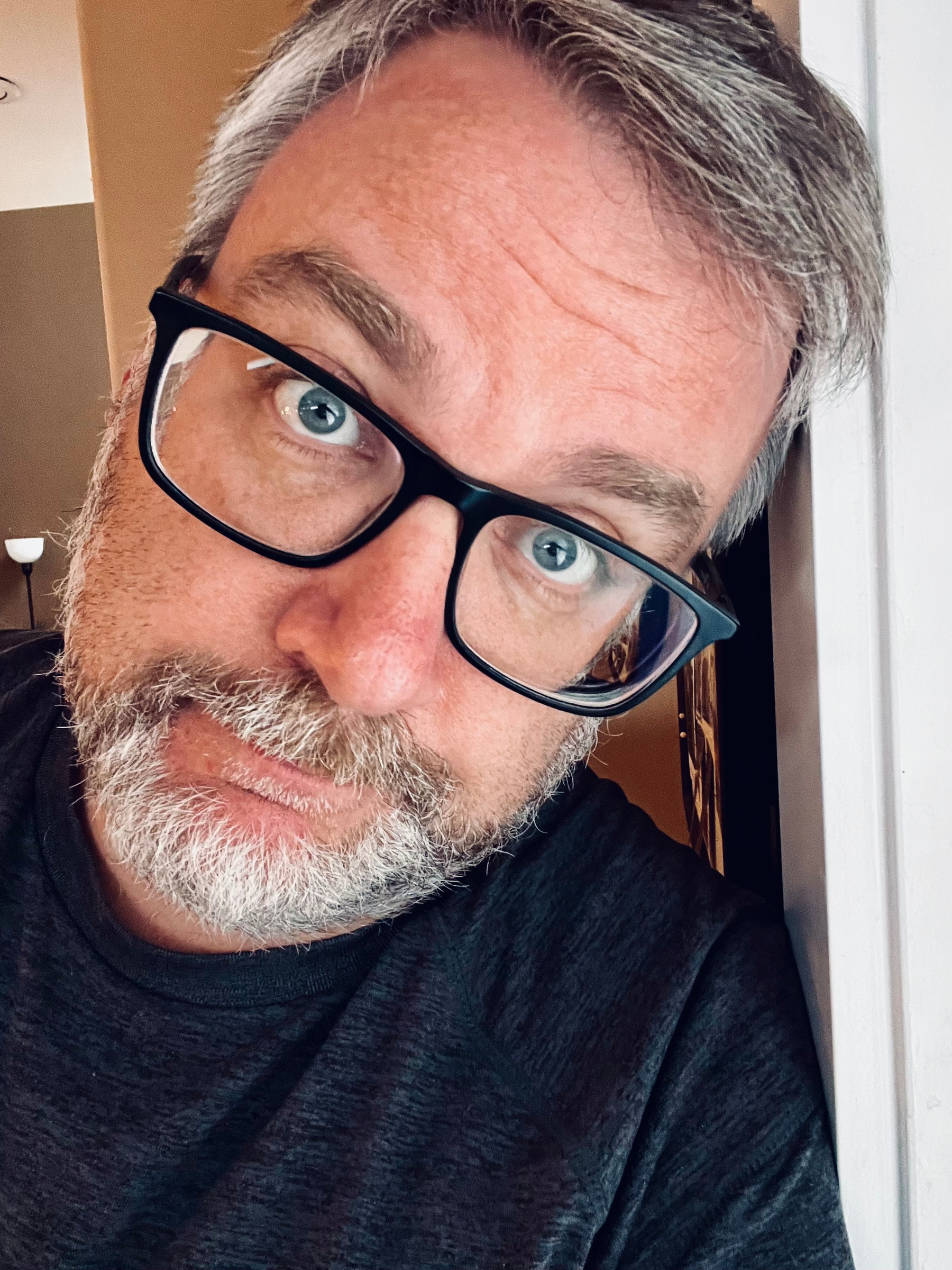 A man with glasses and a beard is leaning against a light-colored surface, looking at the camera.