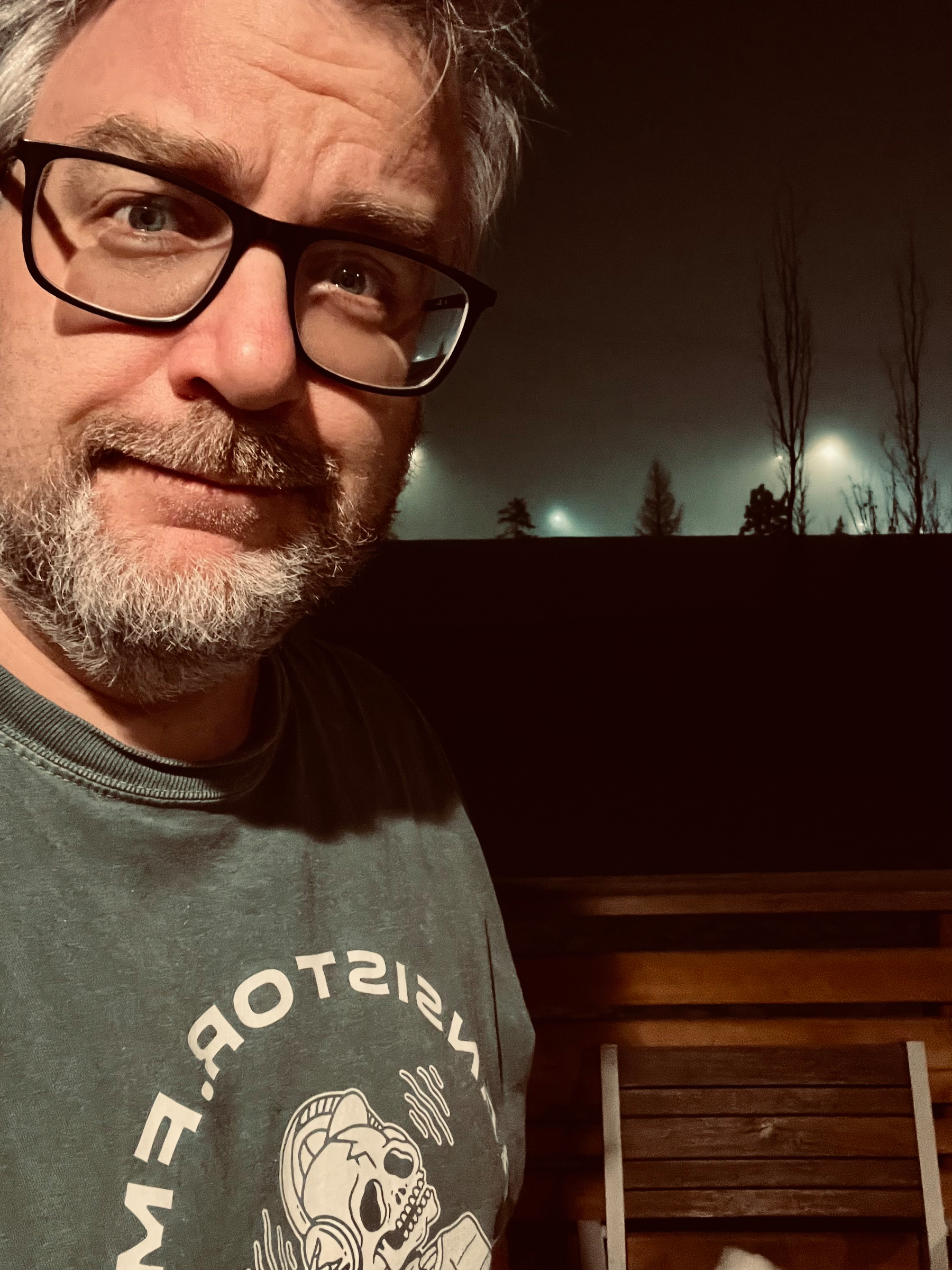 A person wearing glasses and a T-shirt stands outdoors at night, with lights and trees in the background.