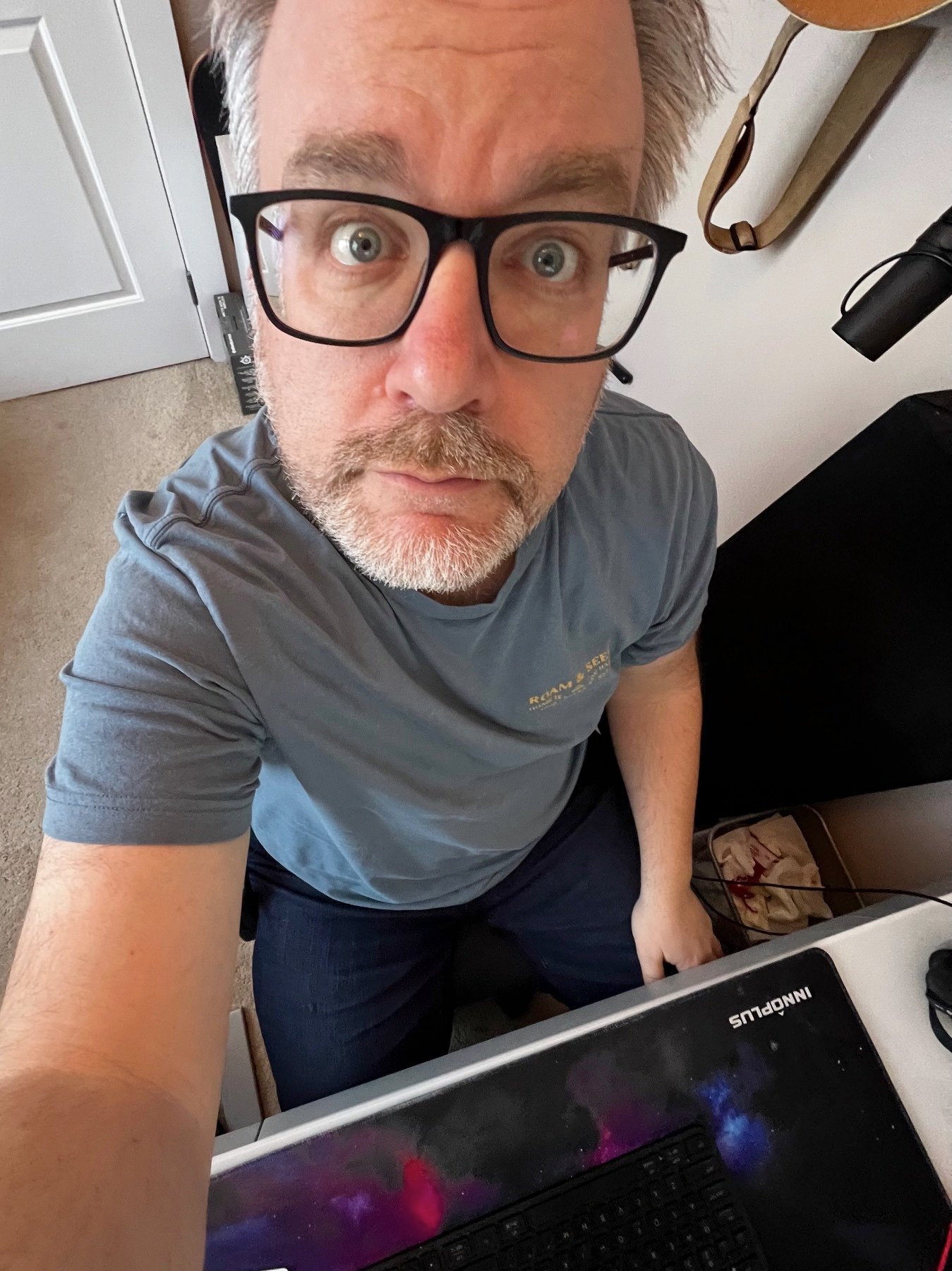 A person wearing glasses and a blue shirt is looking up at the camera while seated near a keyboard and mouse pad.