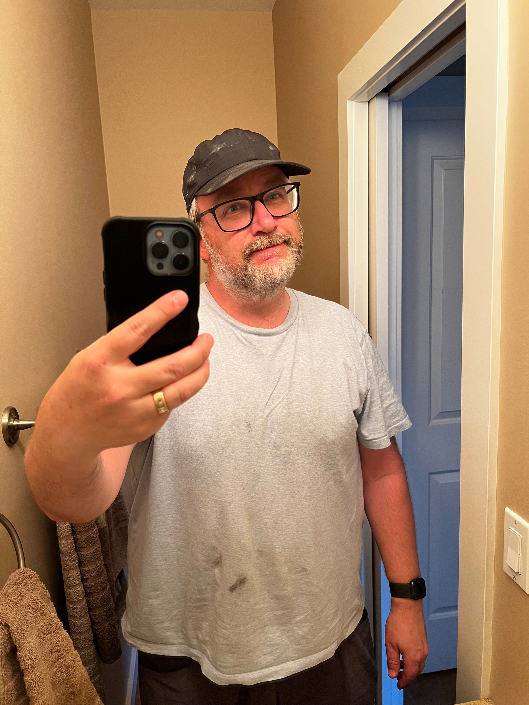 A bearded man wearing glasses and a cap stands in front of a mirror, taking a selfie with a smartphone in a bathroom.