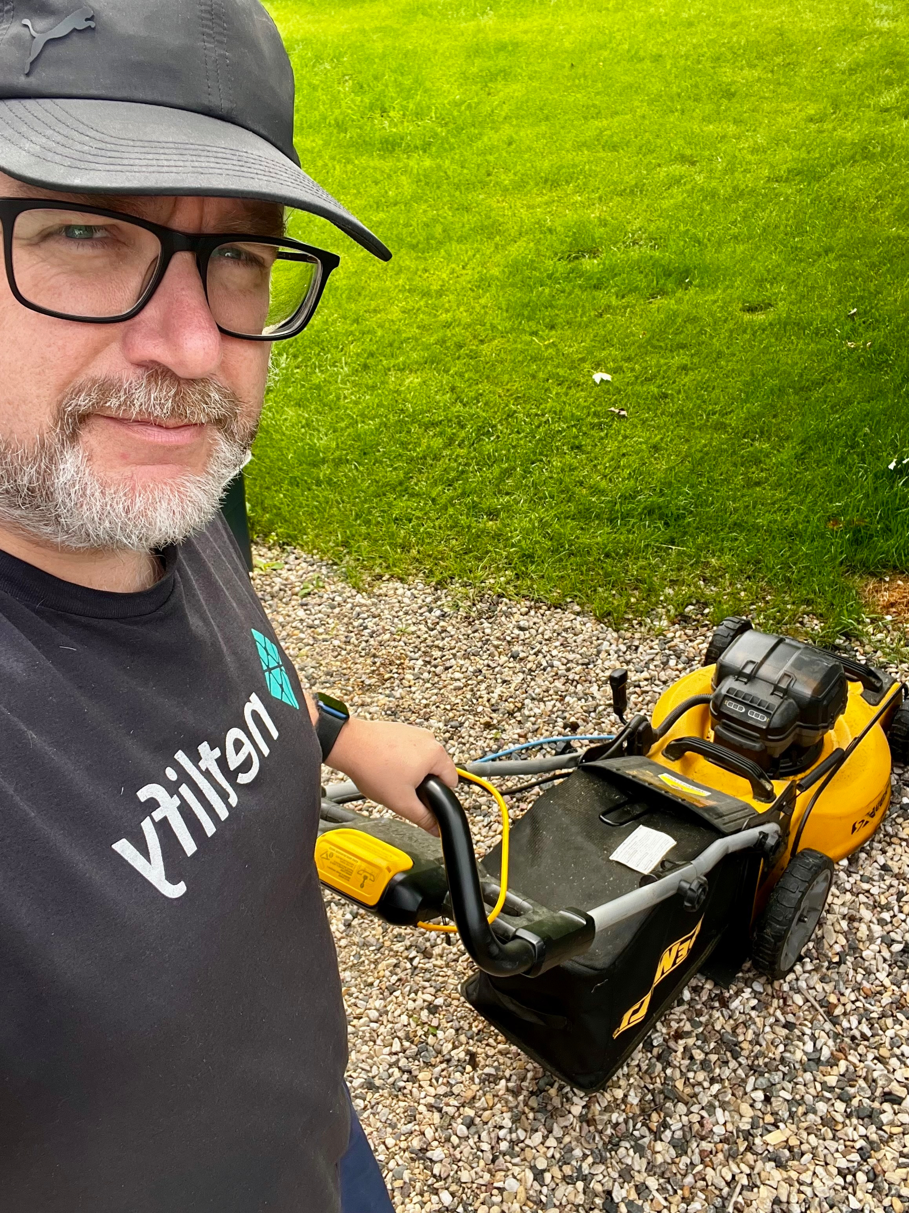 A man wearing glasses and a cap is standing on a gravel path holding a yellow lawn mower with a grassy lawn in the background.