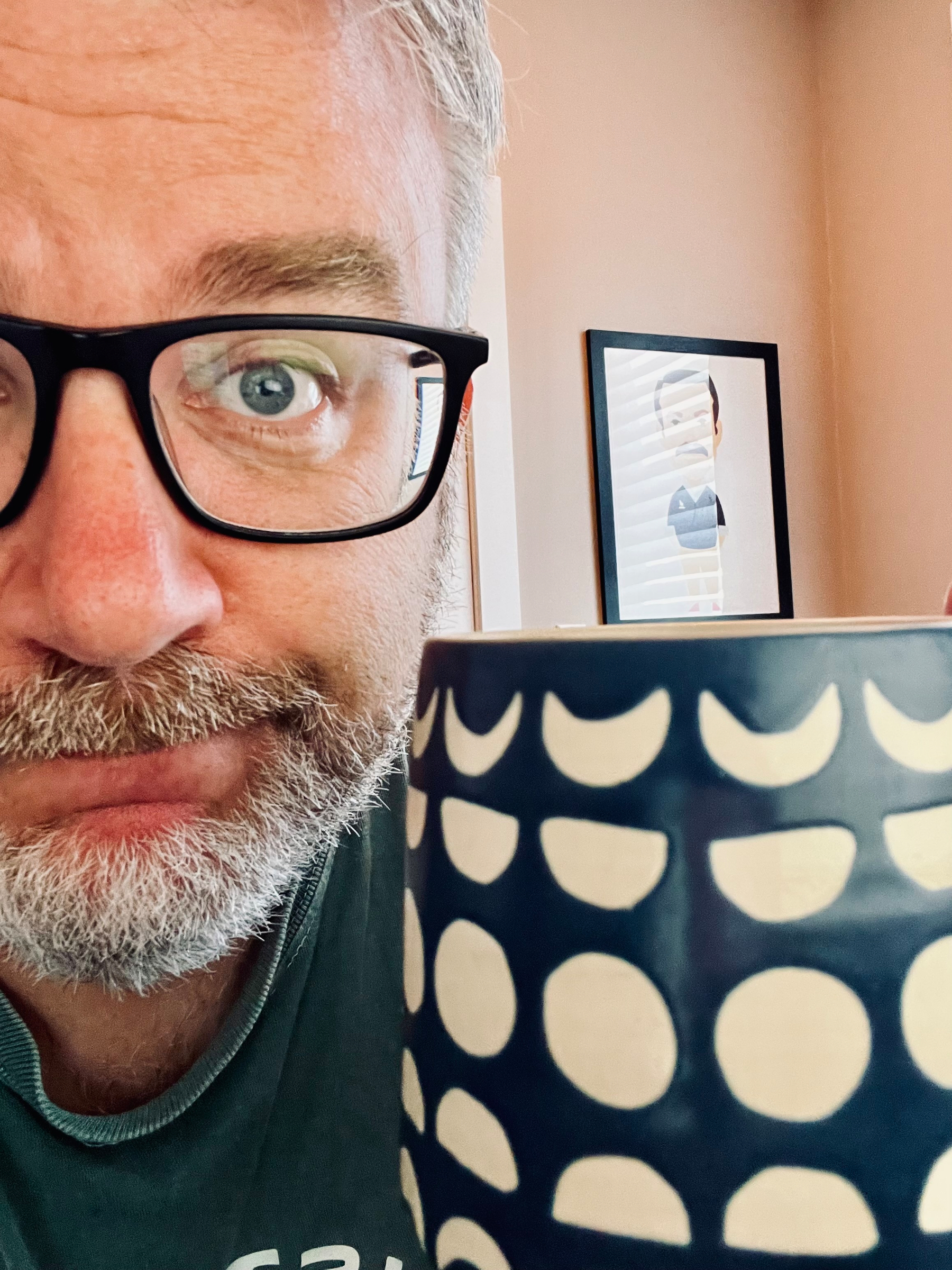 A man with gray hair and glasses is holding a dark blue cup with white patterns close to the camera, with a framed picture of Ted Lasso visible in the background.