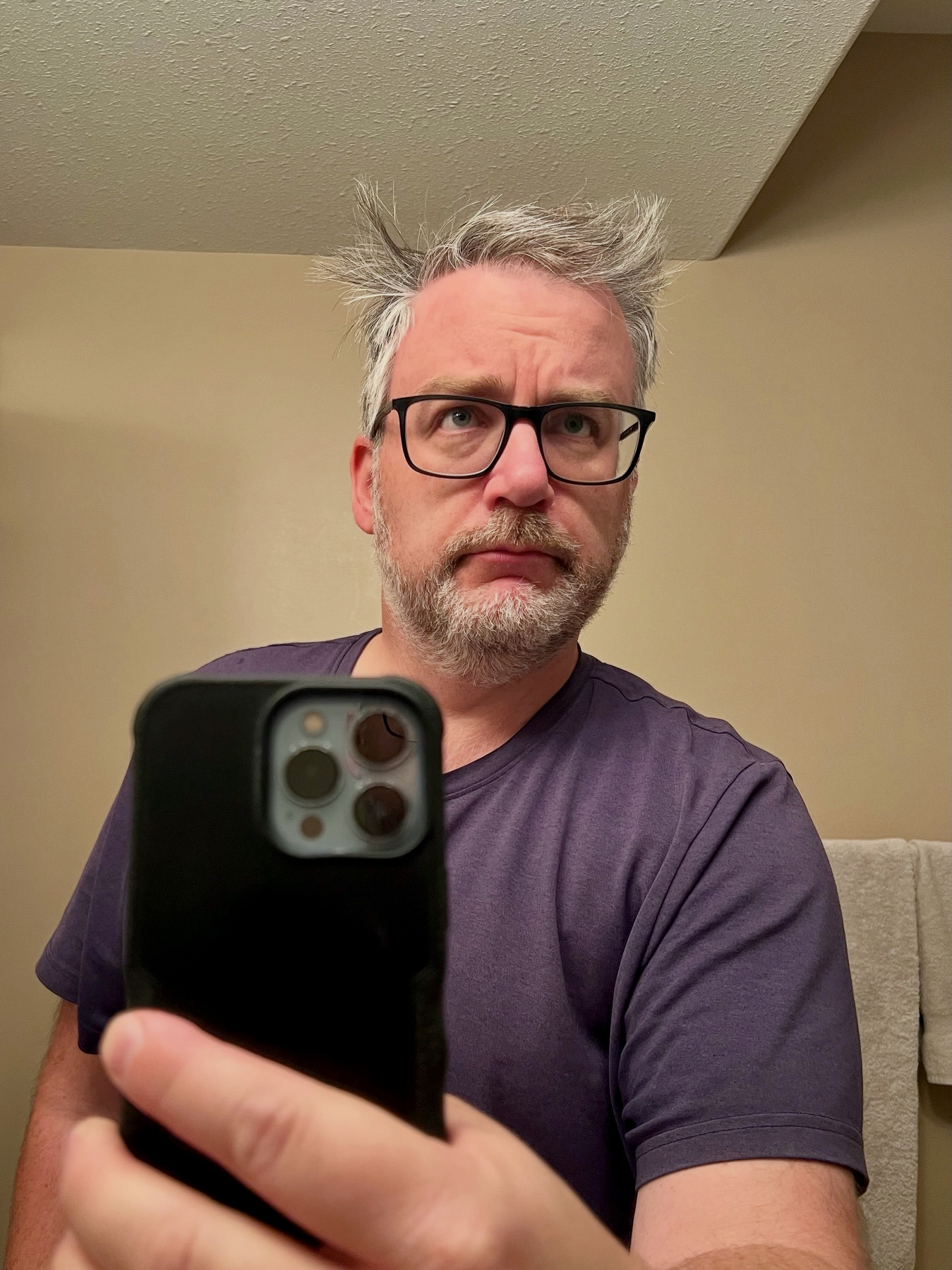 Mirror Selfie of me looking in the mirror with hair askew in multiple directions. iPhone in the foreground and me in the background.