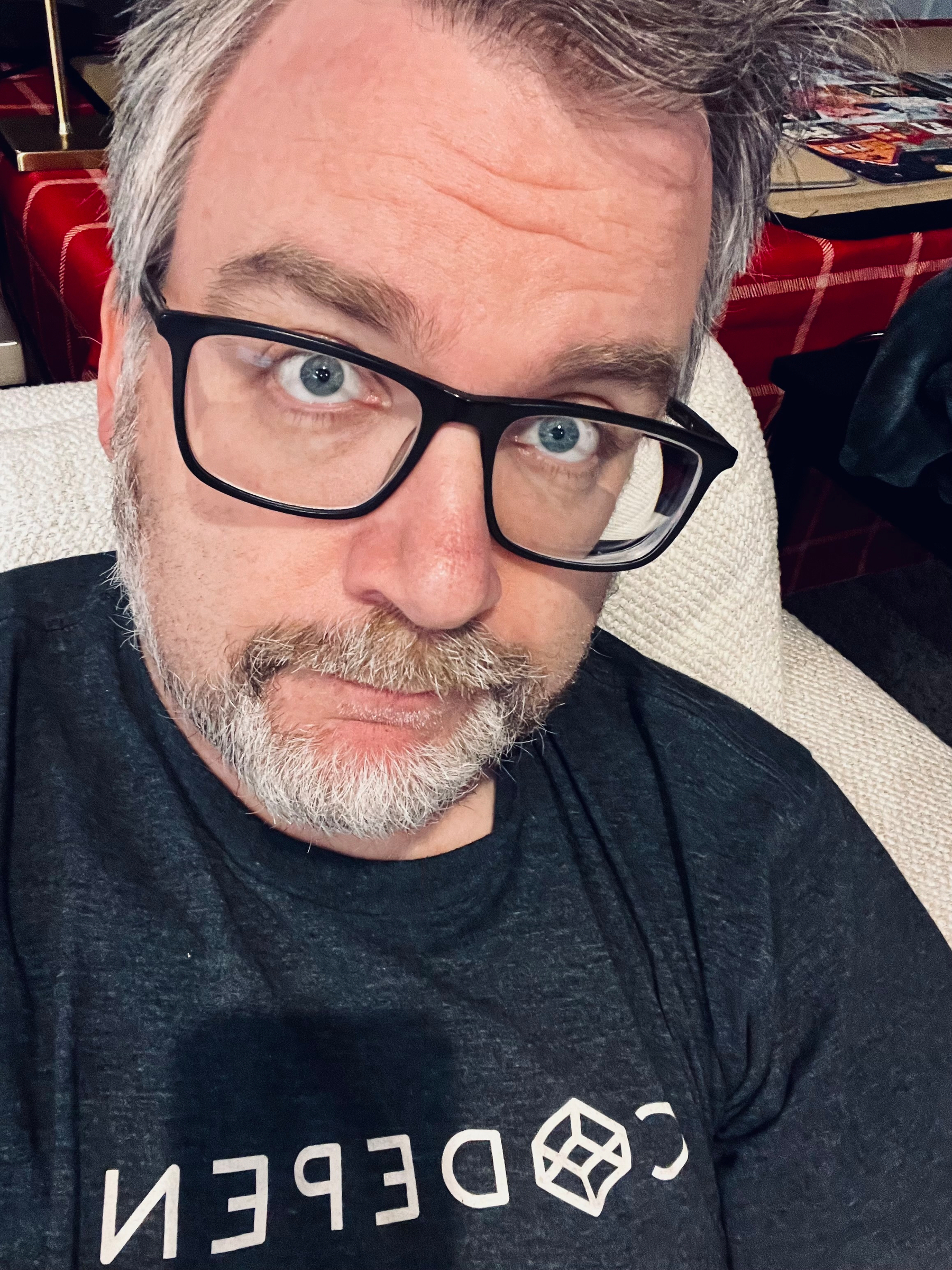 A man with glasses and a beard is looking at the camera while wearing a dark t-shirt with text on it.