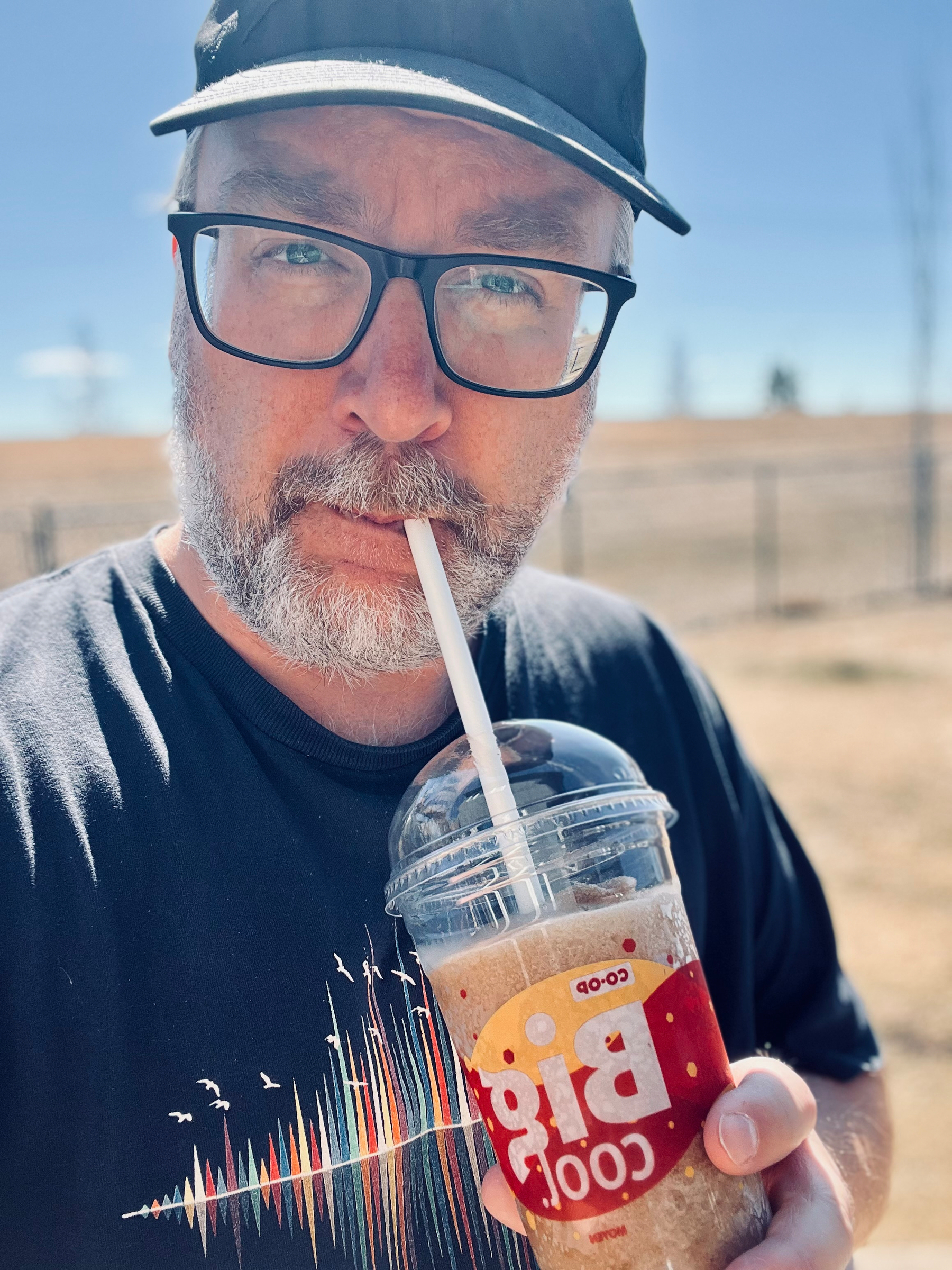 A bearded man wearing glasses and a cap drinks from a cup with the label Big Cool outdoors.