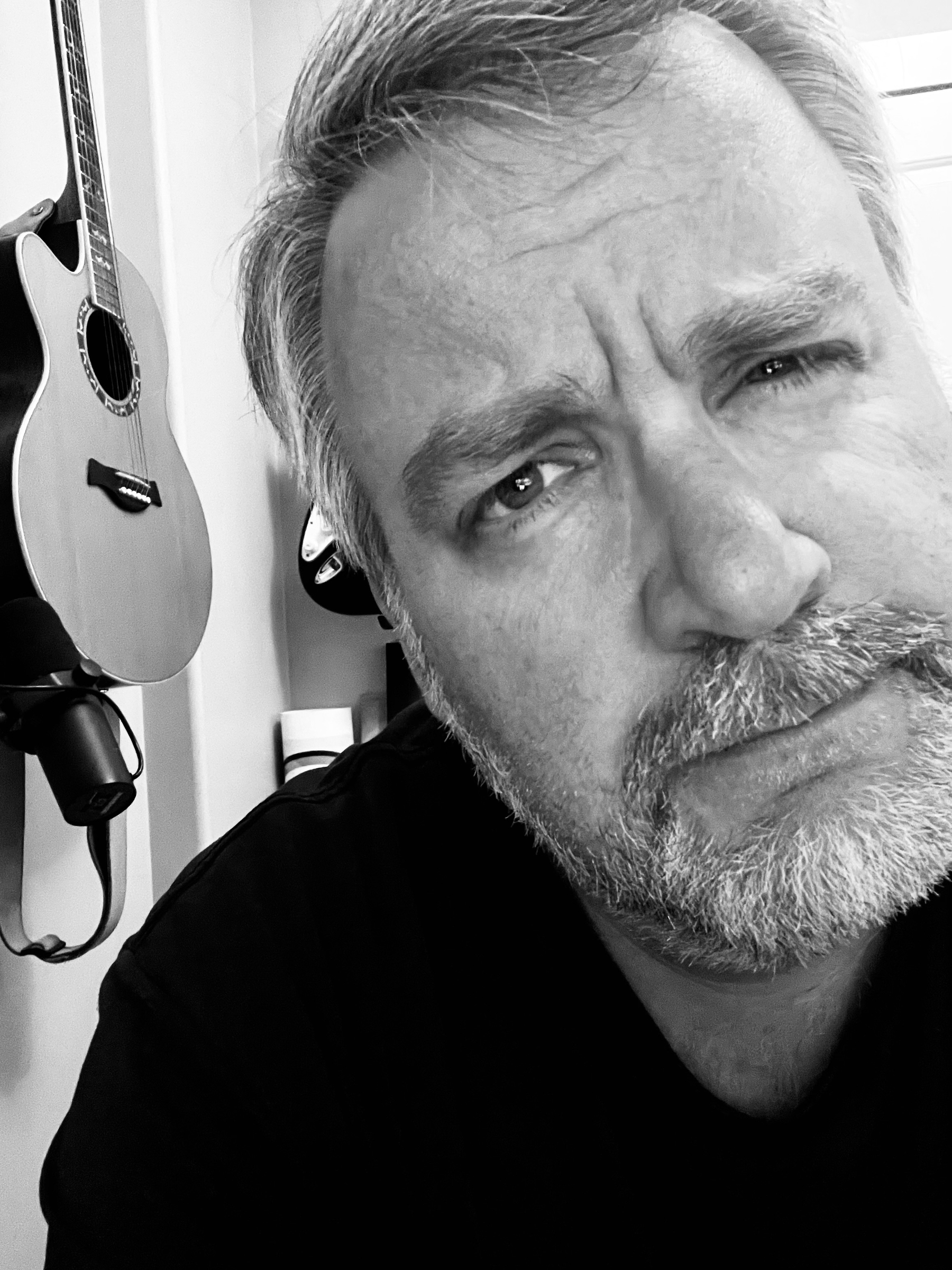 A bearded person looks into the camera with a slightly furrowed brow, while guitars hang on the wall in the background.