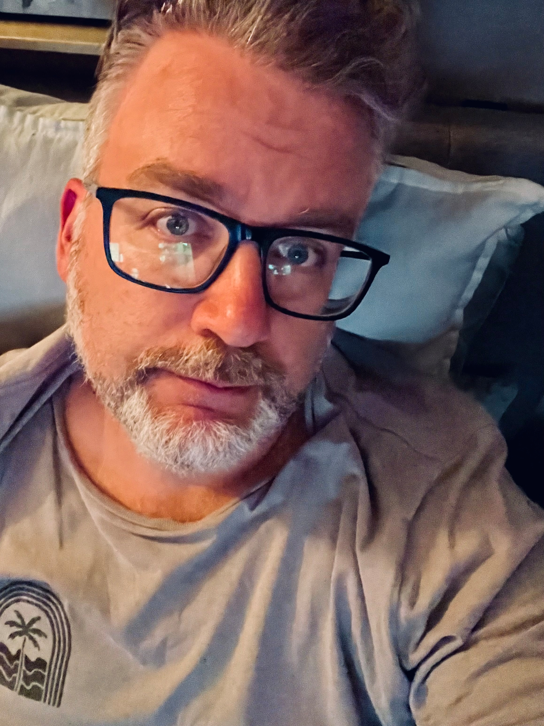 A middle-aged man with gray hair and beard, wearing glasses and a casual shirt, is reclining on a pillow with a relaxed expression.