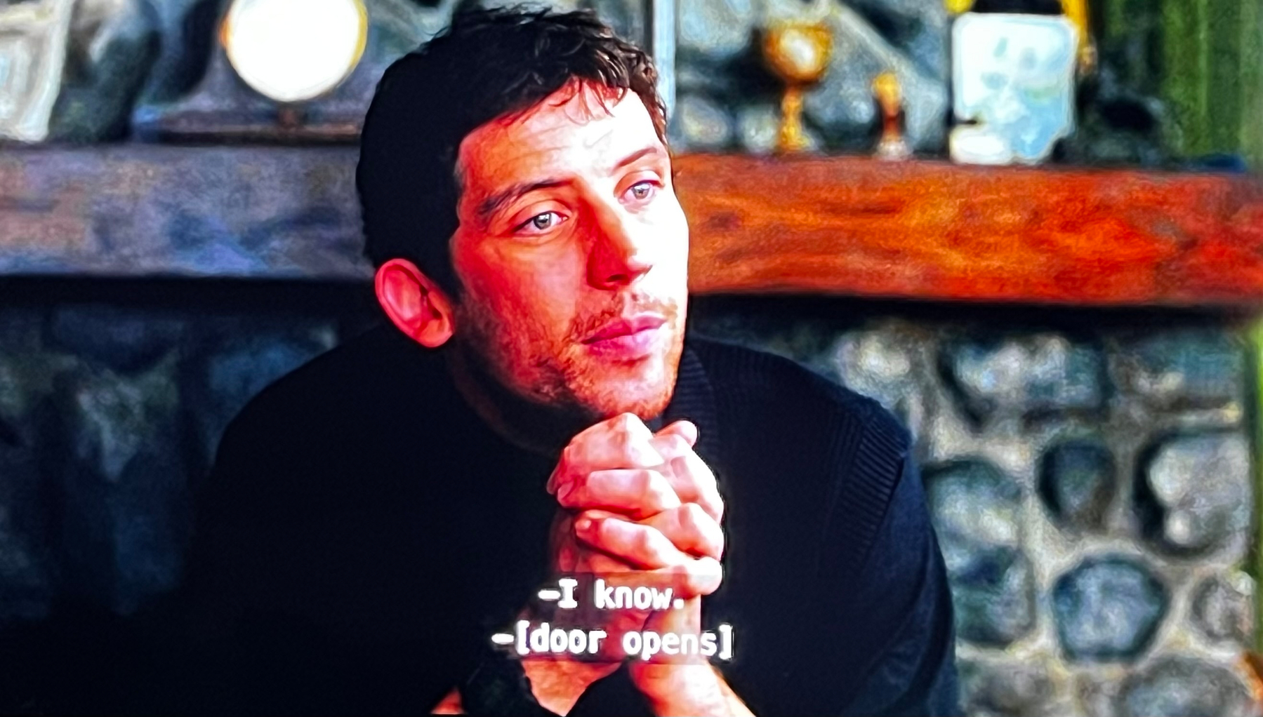 A man is sitting with clasped hands, looking to the side, with subtitles reading -I know. -[door opens].