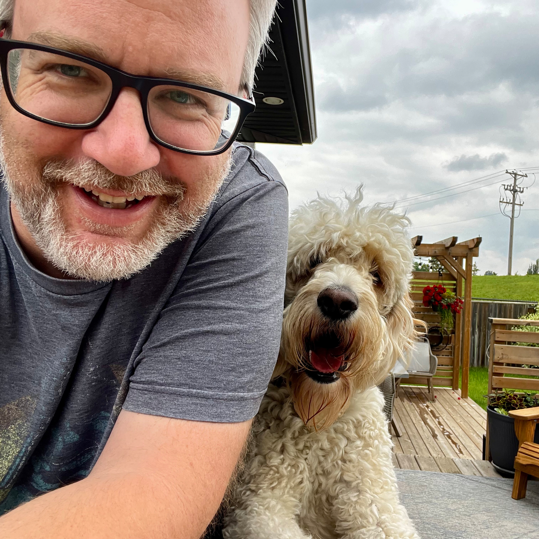 A man wearing glasses and a gray shirt is smiling next to a fluffy white dog with curly hair outdoors, with a wooden deck and garden visible in the background.