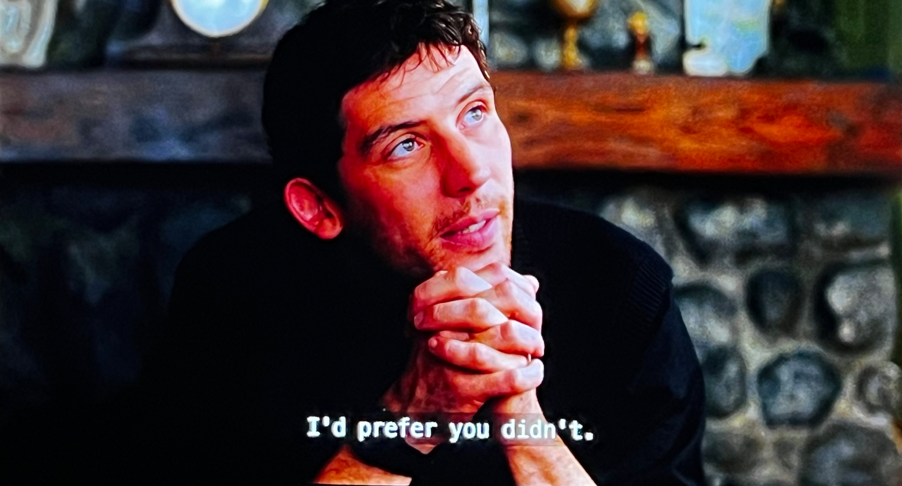 A person sits with clasped hands, looking upward, with subtitles reading I'd prefer you didn't.