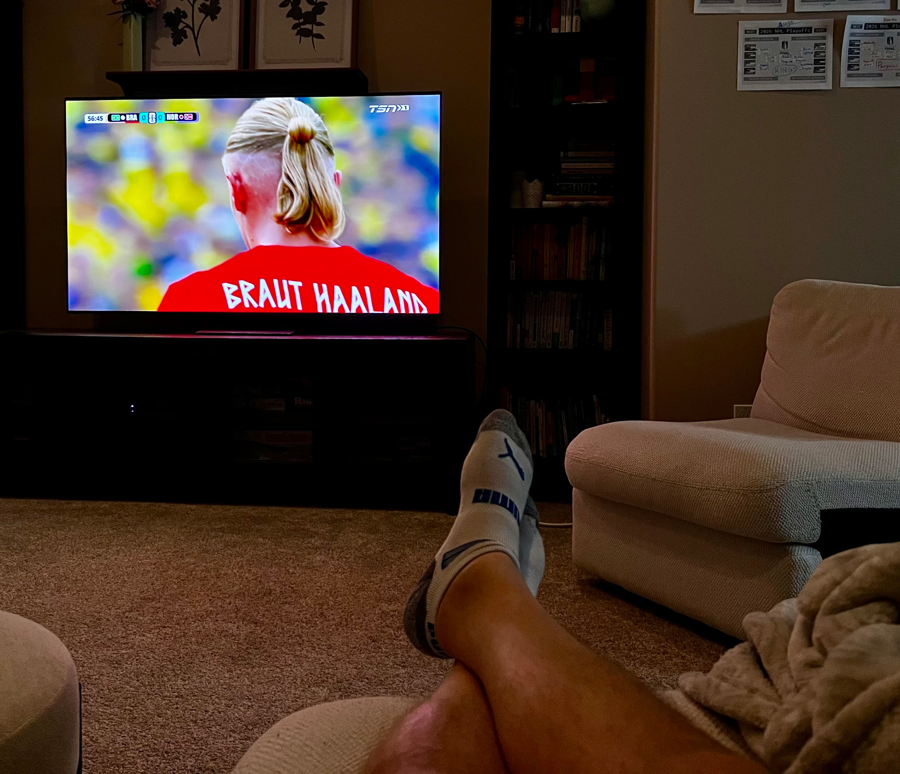 A person is relaxing with their feet up while watching a soccer match featuring a player named Braut Haaland on TV.