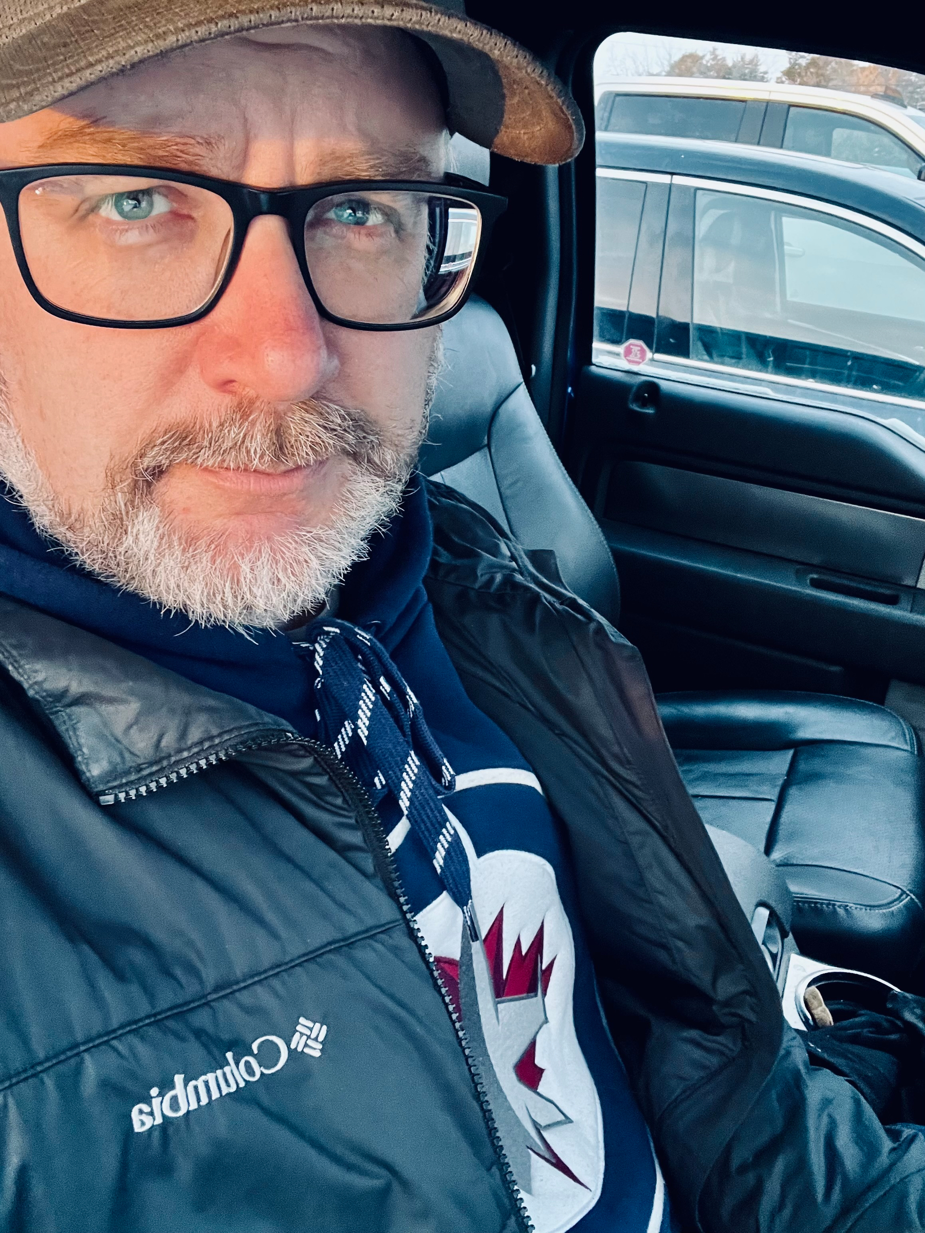 A man with glasses and a beard is sitting in a car, wearing a Columbia jacket and a hoodie with a Winnipeg Jets hockey team logo.