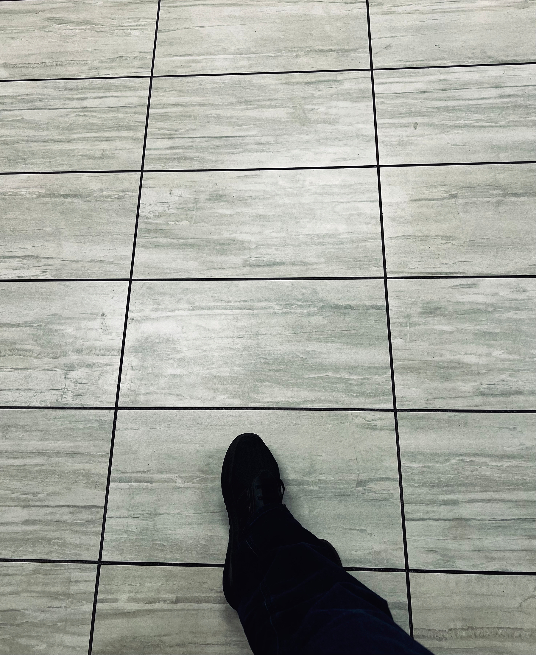 A person wearing a black shoe and dark pants is standing on a tiled floor with light gray, marble-like patterns.