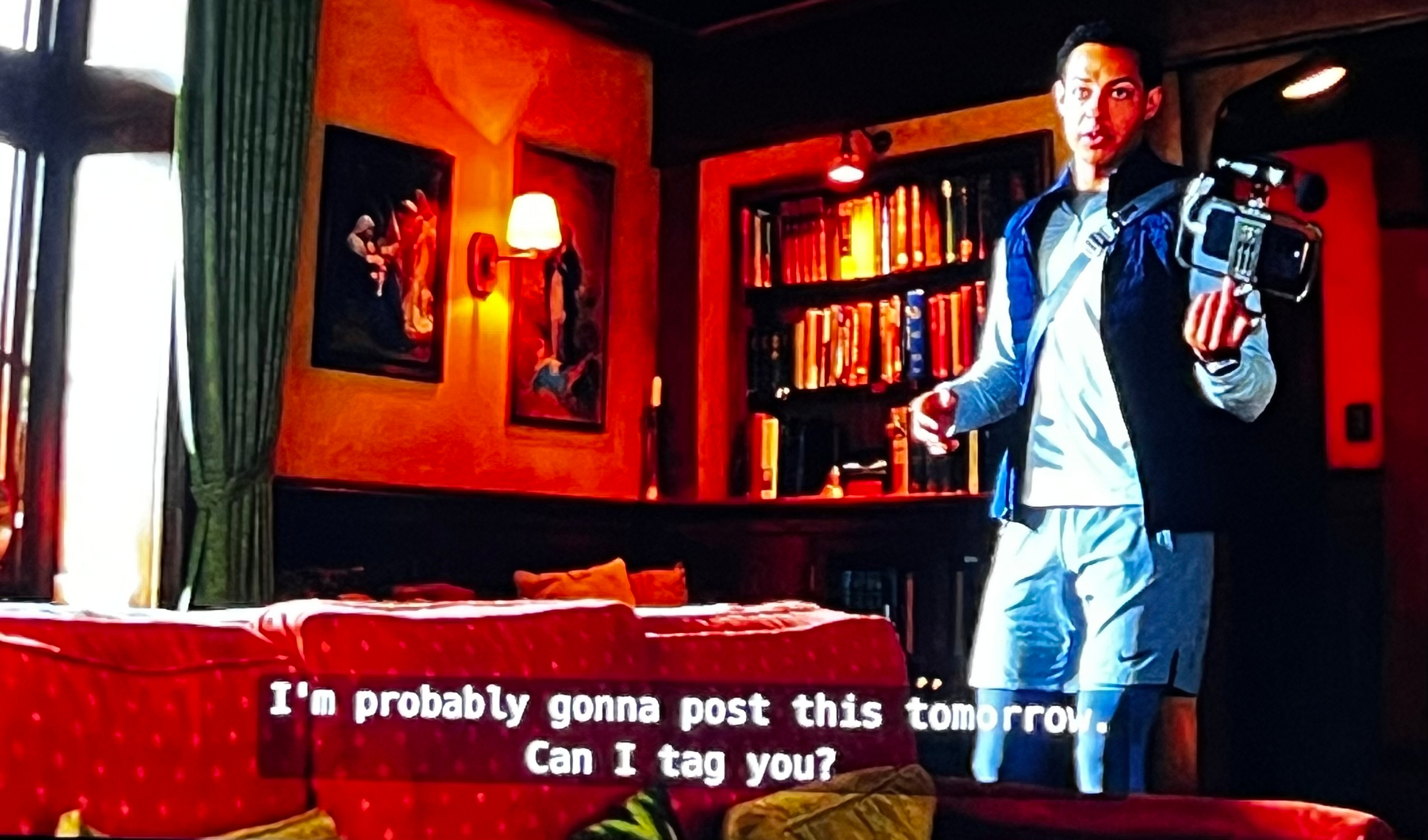 A person holding a camera stands in a cozy room featuring a sofa, bookshelves, and paintings, with subtitles saying, I'm probably gonna post this tomorrow. Can I tag you?