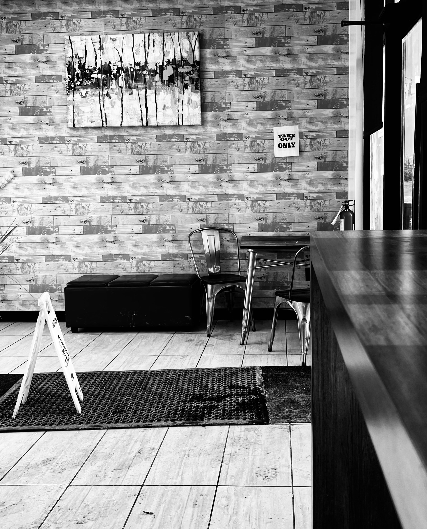 A monochromatic café interior features a wooden wall with contemporary art, metal chairs, and a Take Out Only sign.