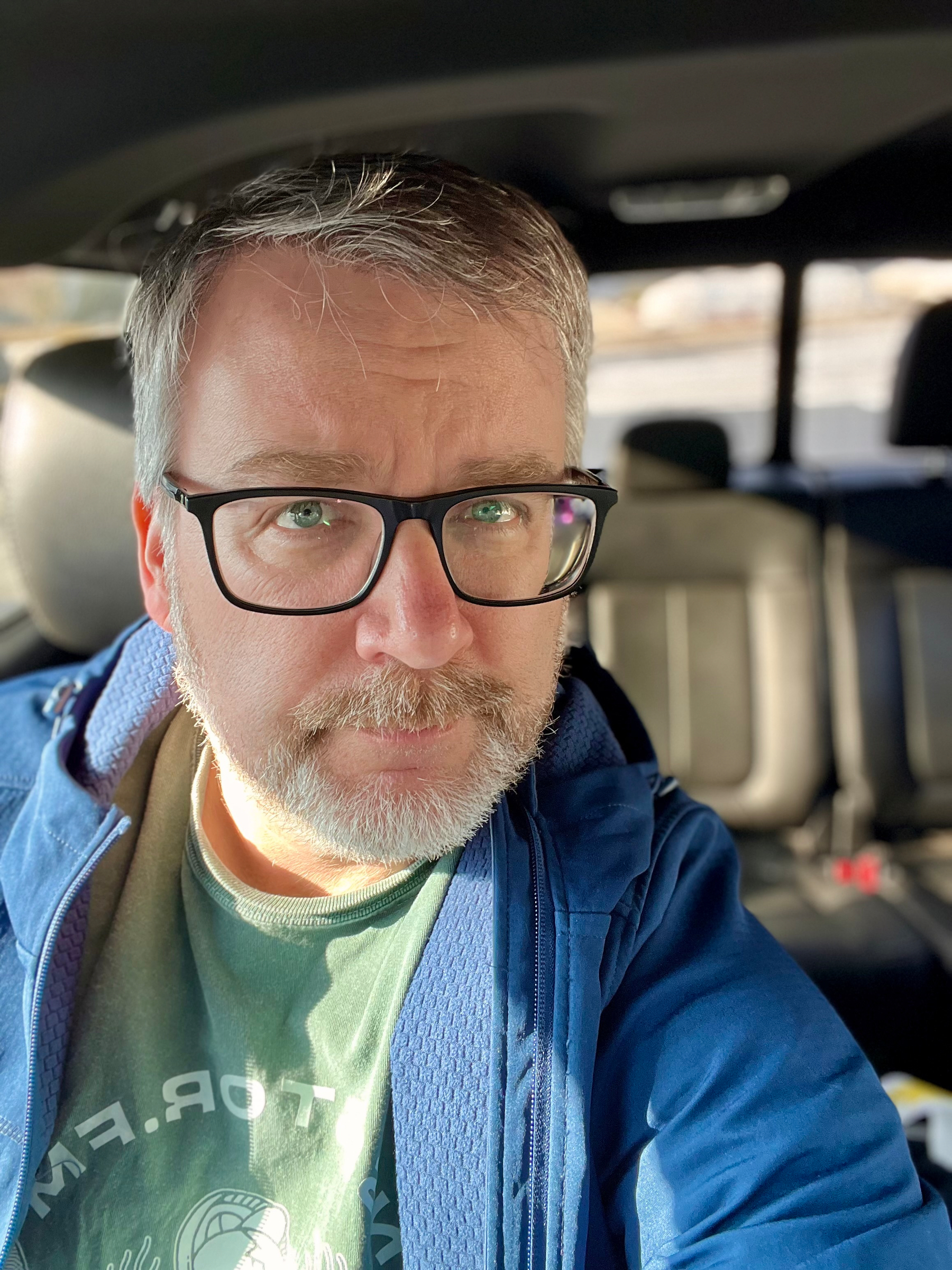 A person with glasses and a beard is sitting in a car, wearing a blue jacket and a green shirt.