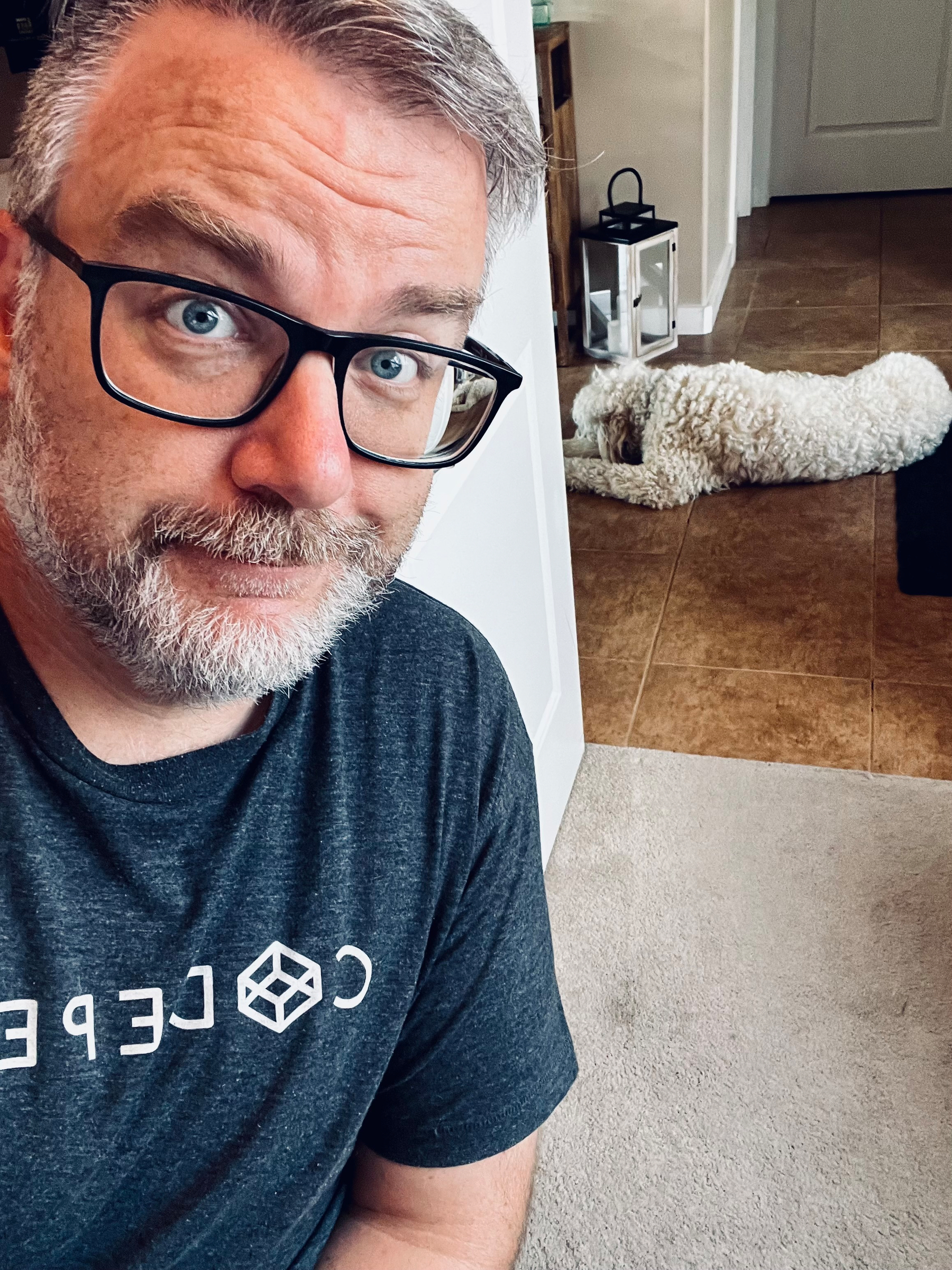 A man with glasses and a beard is taking a selfie indoors while a white dog lies on the floor in the background.