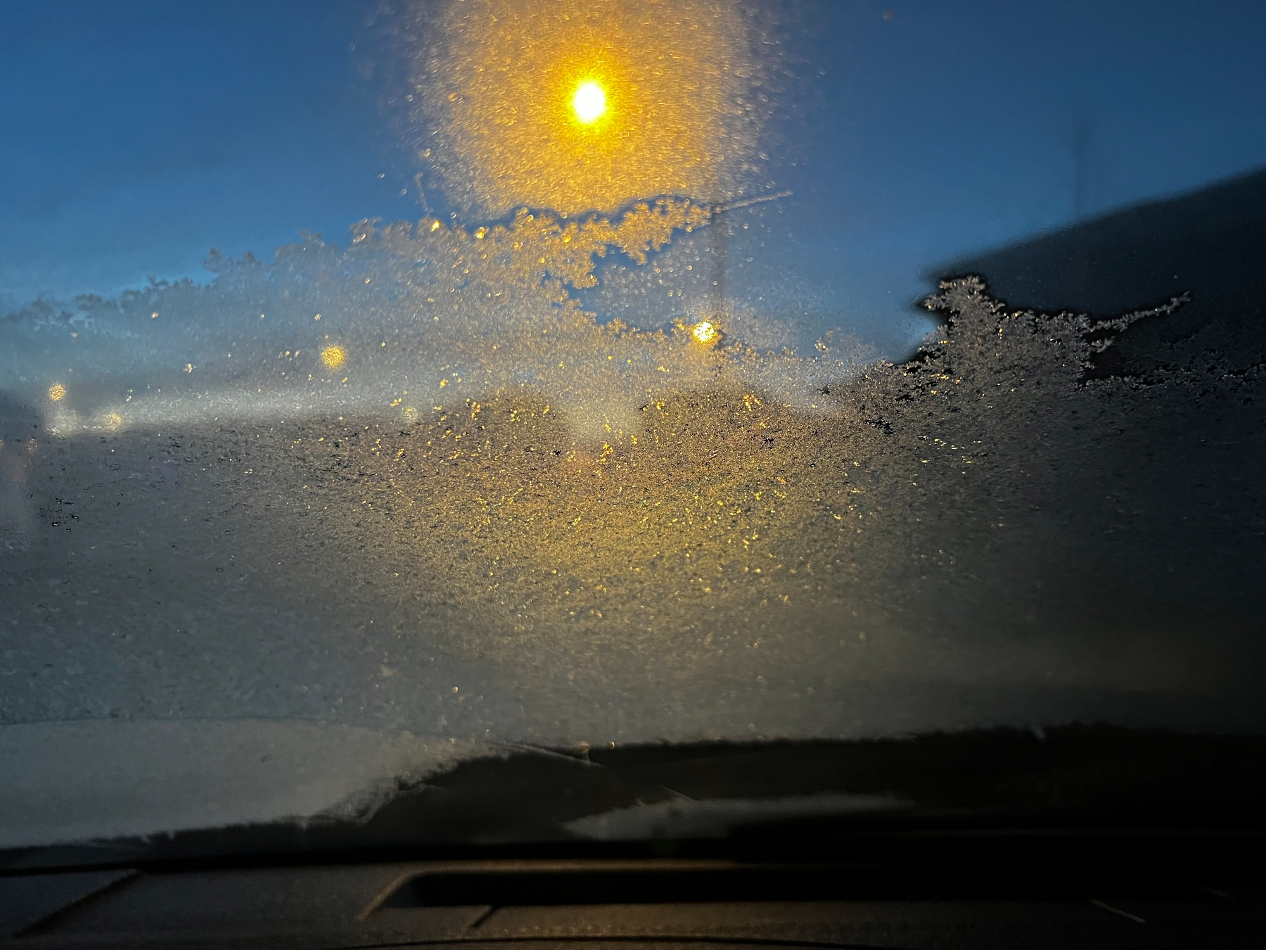 Frost partially covers a car windshield, with streetlights glowing through the icy patterns during early morning or evening.