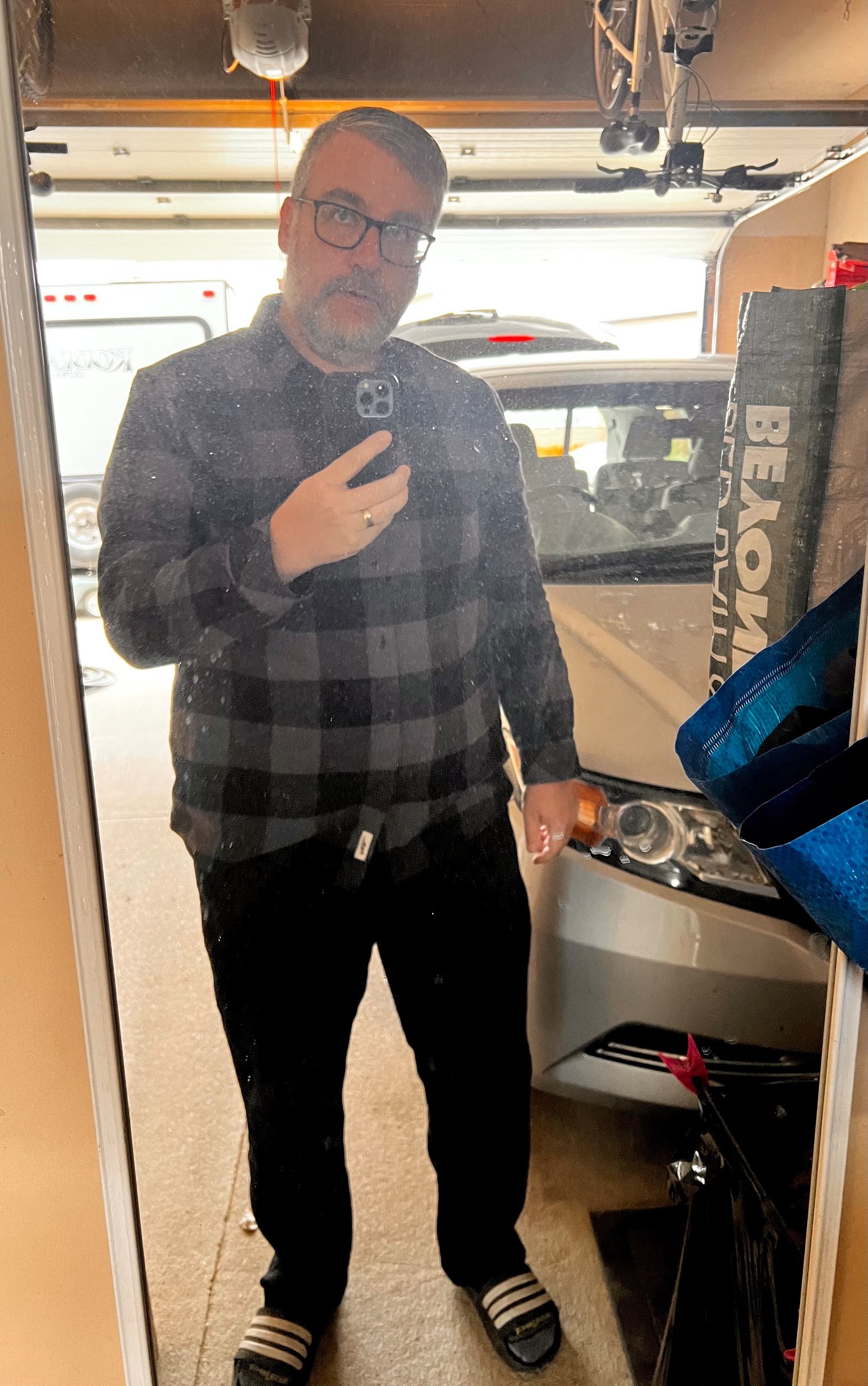 A man wearing glasses, a black and gray checkered shirt, black pants, and striped slippers is taking a mirror selfie in a garage with a car and various items visible in the background.