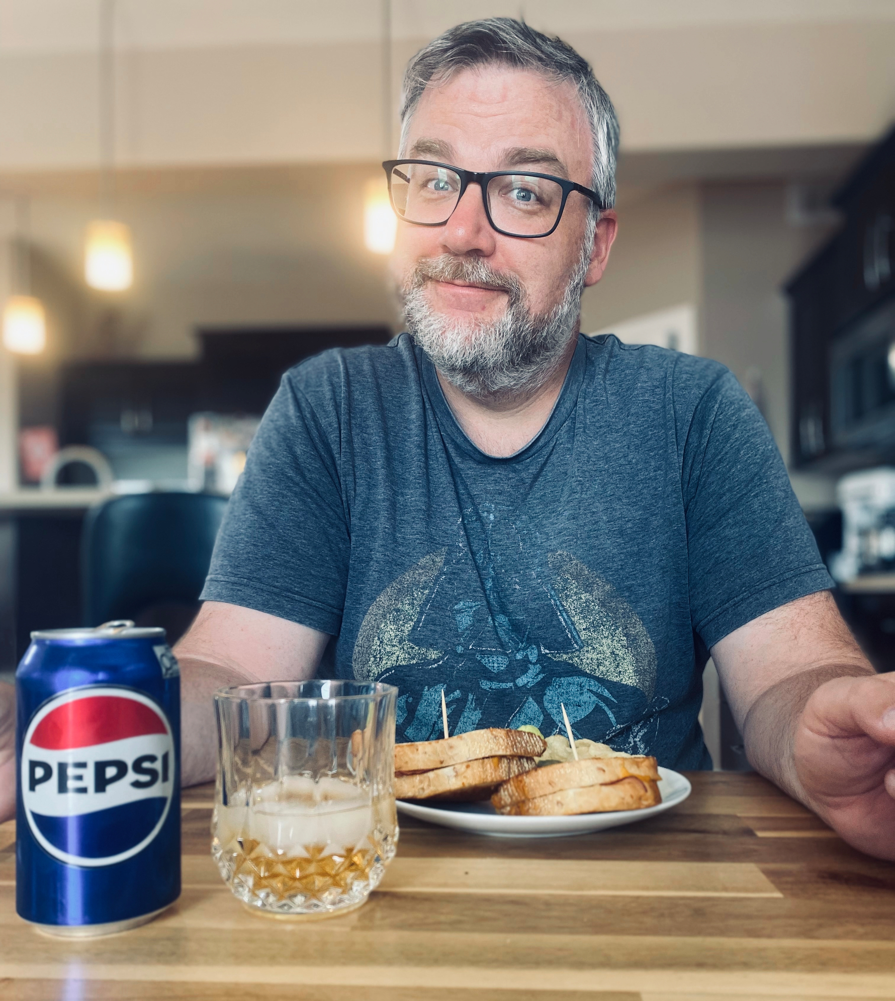 A man wearing glasses and a graphic t-shirt is sitting at a table with a plate of sandwiches, a glass of drink, and a can of Pepsi.