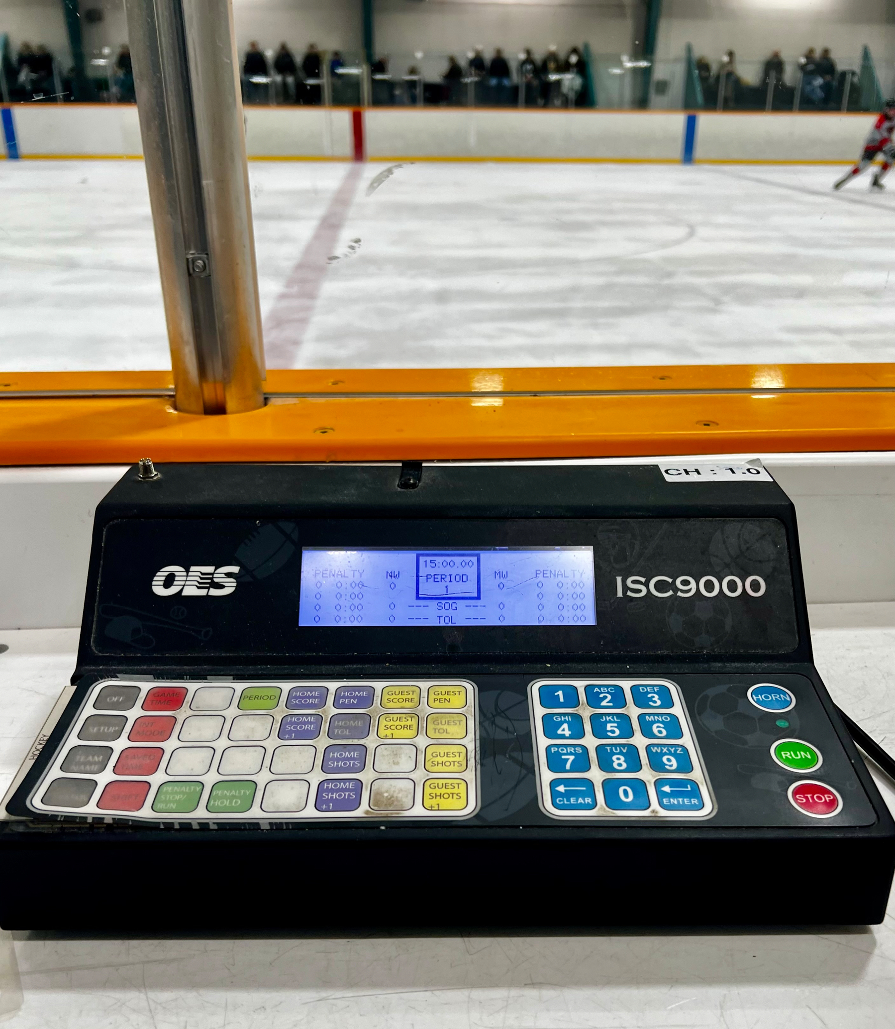 A control panel for an ISC9000 scoreboard is set up at an ice hockey rink, with a game in progress in the background.