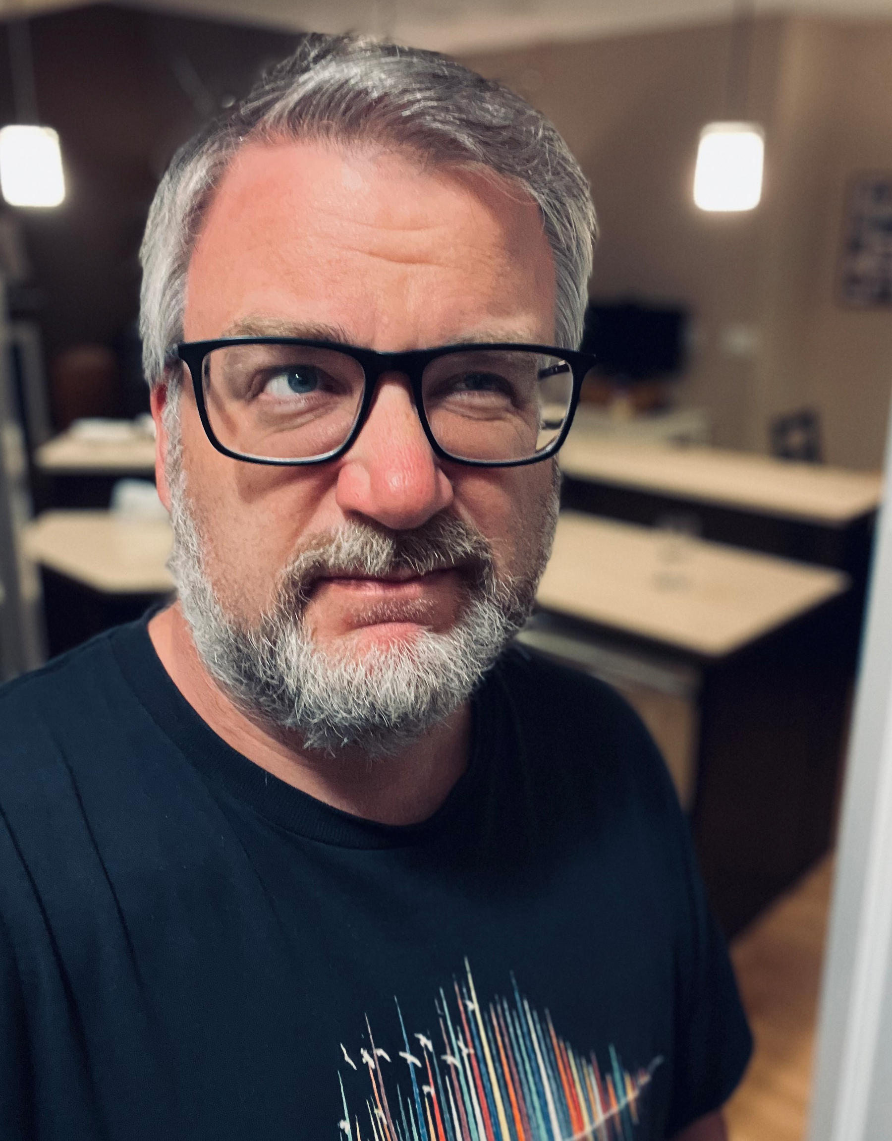 A man with gray hair, a beard, and black-rimmed glasses is looking slightly to the side while wearing a black t-shirt, standing in a softly lit indoor setting.