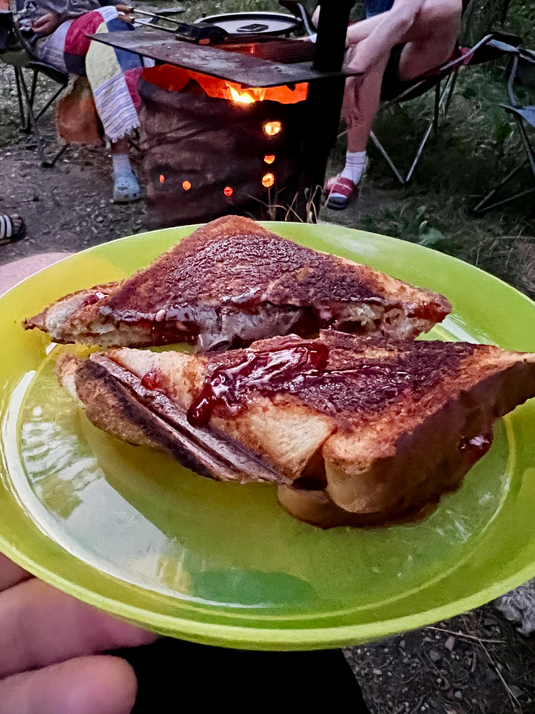 Grilled sandwiches with melted chocolate filling are served on a green plate in an outdoor setting near a campfire with people sitting in camping chairs.