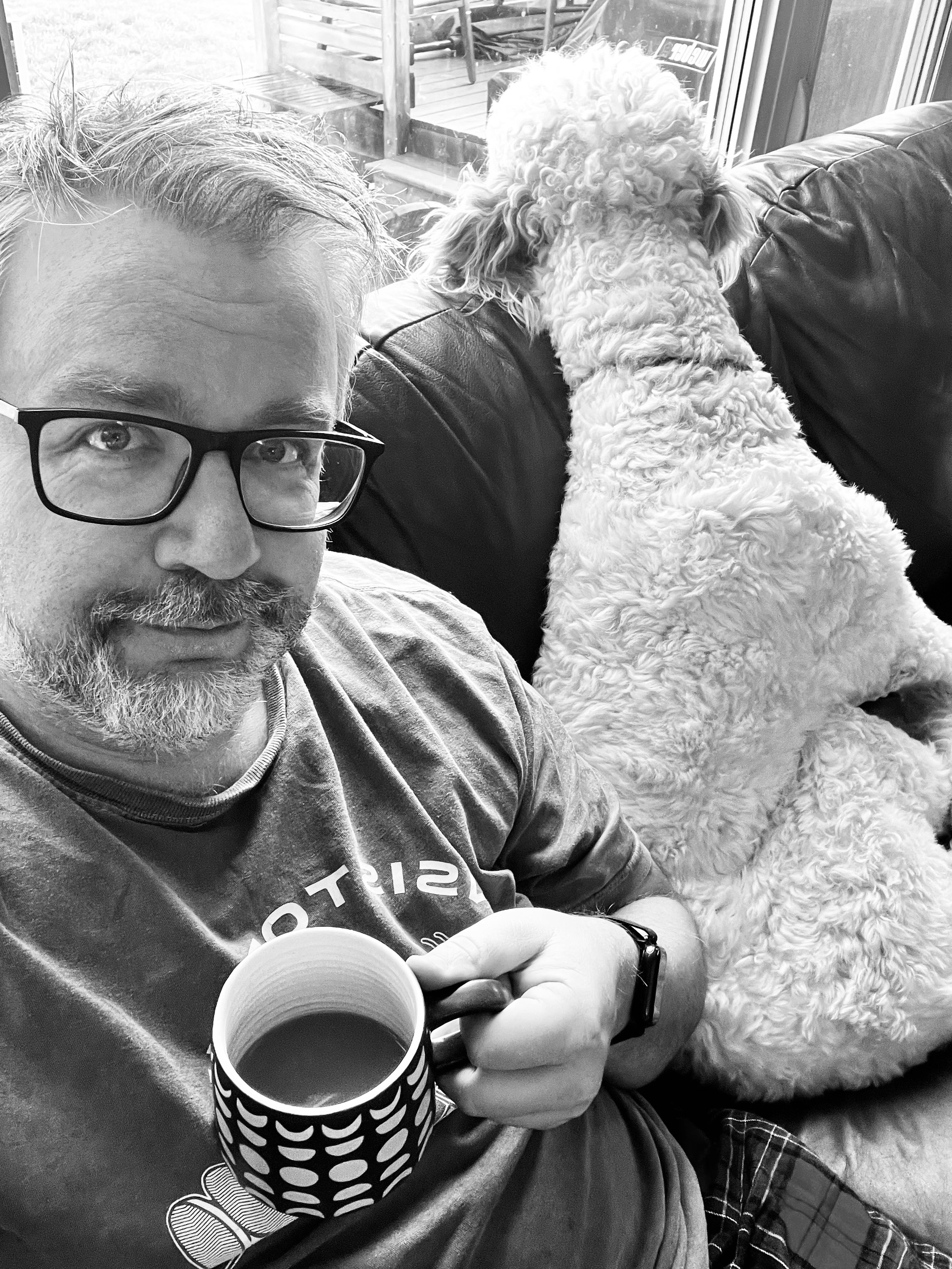 A man holding a mug of coffee sits on a couch next to a large, curly-haired dog who is looking away.